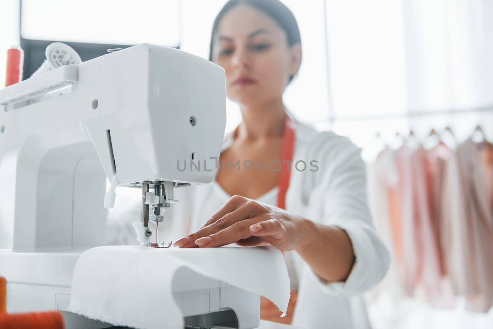 Working by using sewing machine. Seamstress is in her office that is with different clothes.