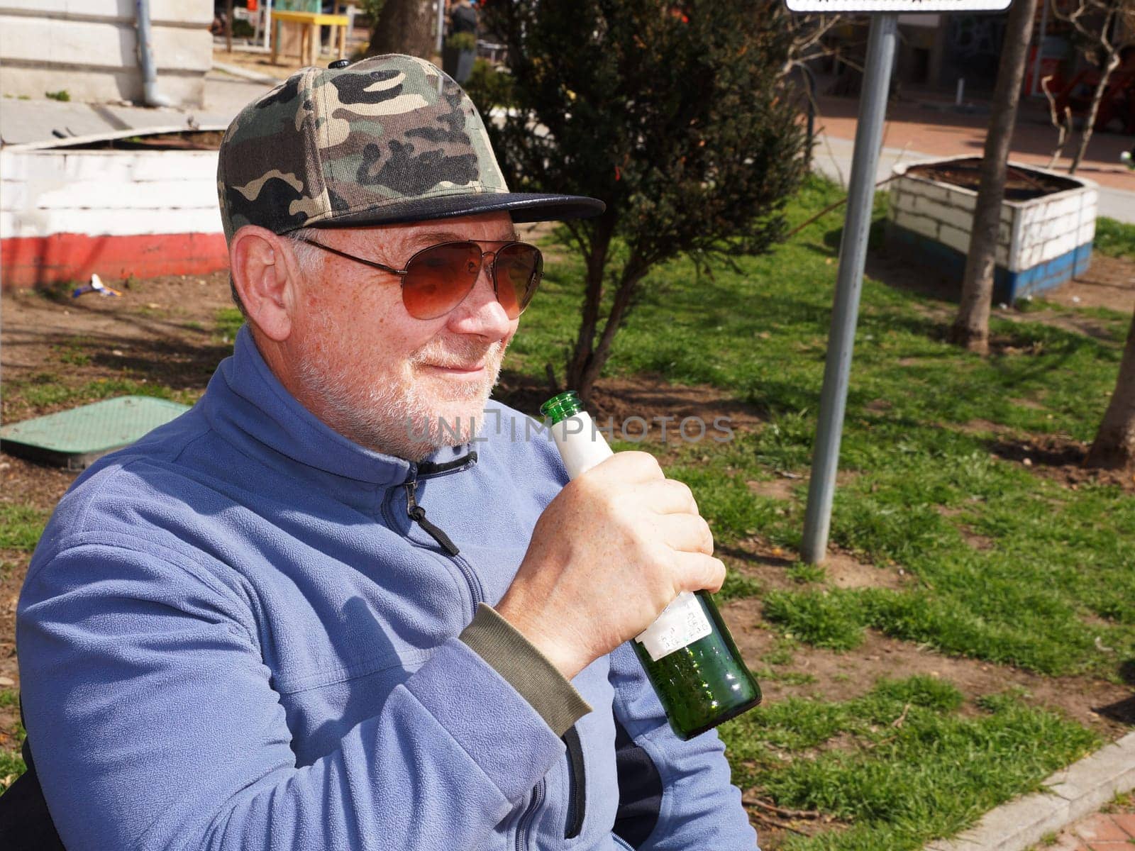 Smiling man in sunglasses drinking beer from bottle in park.