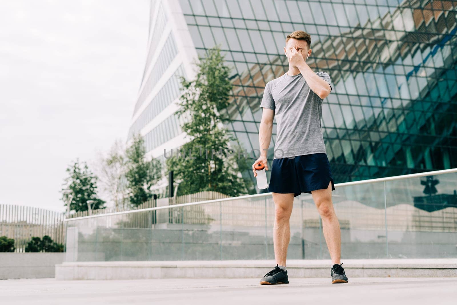 Tired Young man runner drinking water, relaxing after sport training. Holding water bottle while doing fitness workout in summer city urban street, cloudy sky. by Ostanina