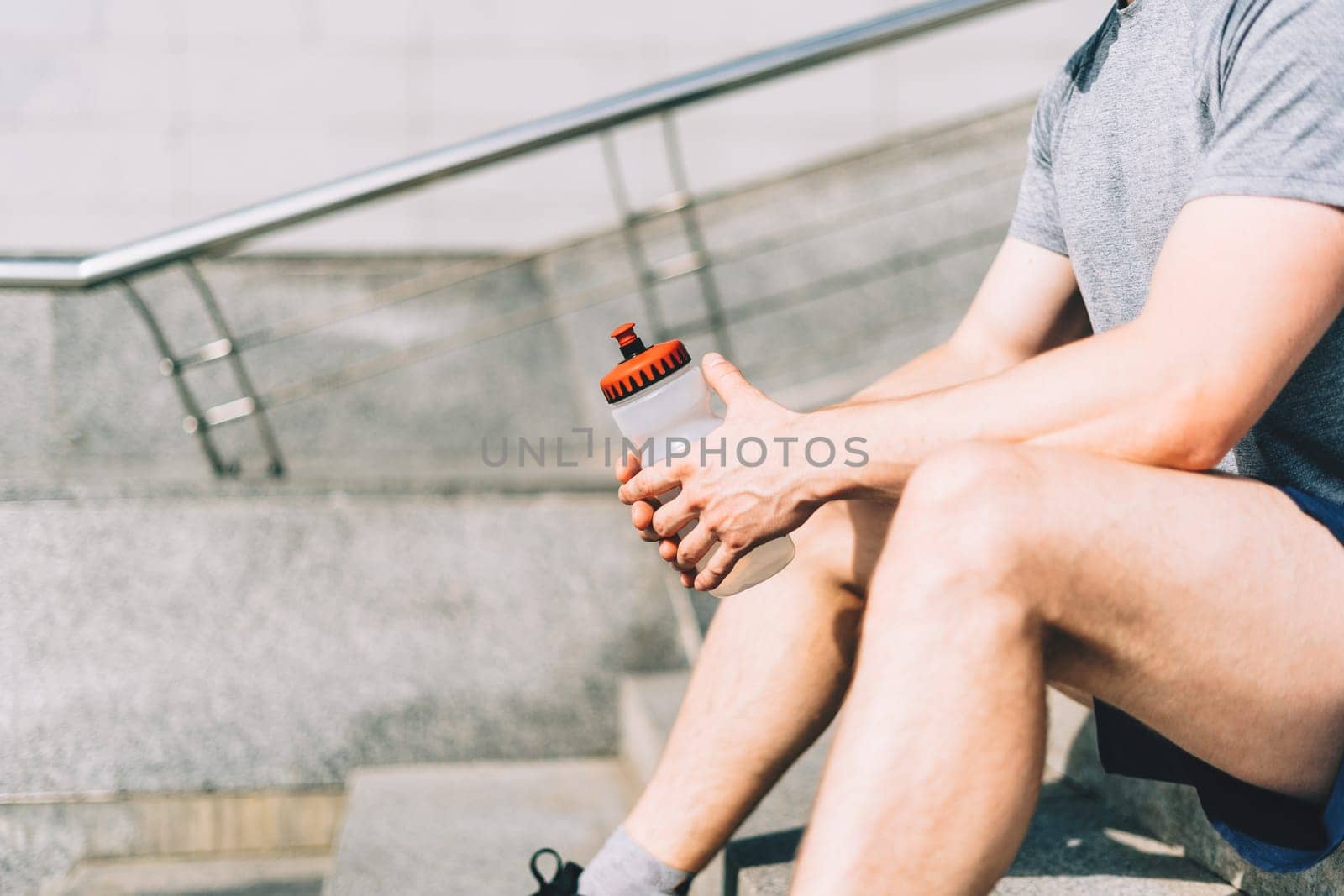 Close up of Tired Young man runner sitting on stairs and relaxing after sport training. Holding water blender bottle while doing workout in summer city street, cloudy sky
