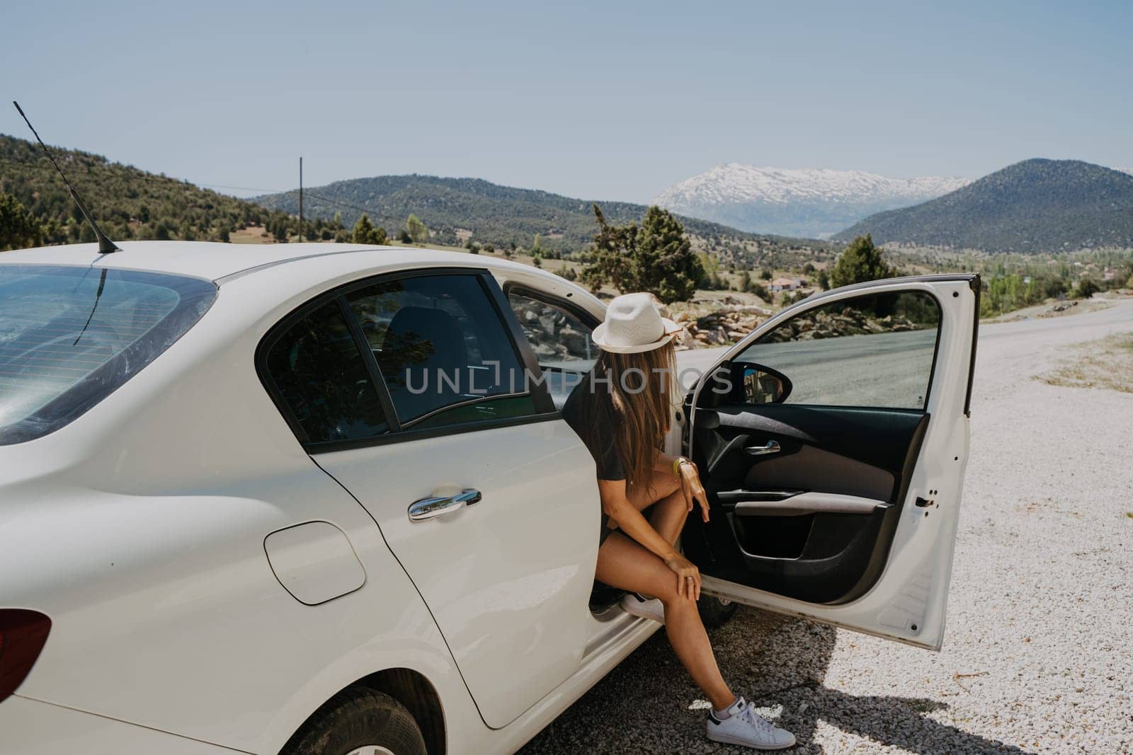 Woman traveler in hat sitting in car with mountain background. Road trip. Young adult woman in sun glasses at summer holiday vacation by Ostanina