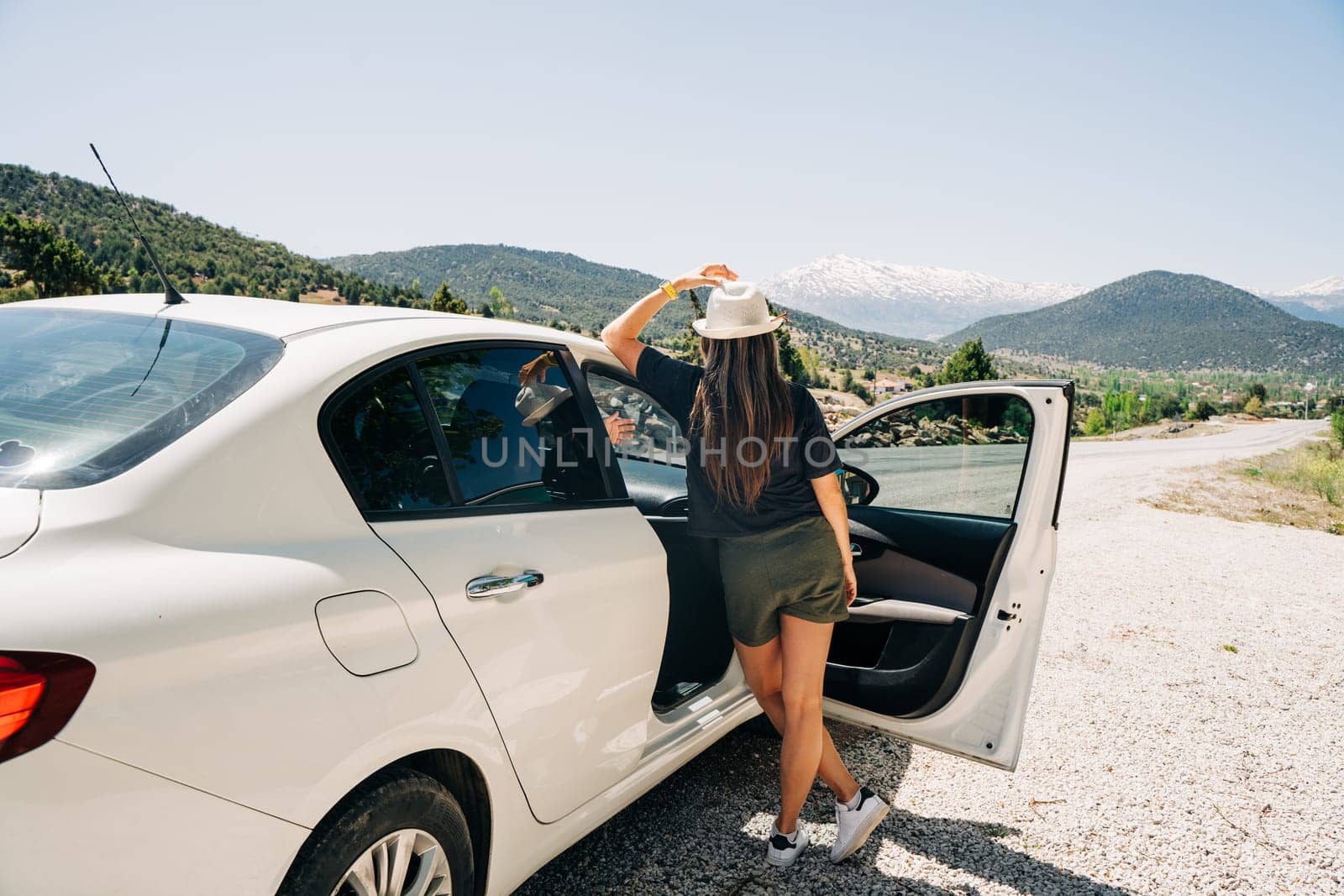 Woman traveler in hat standing in car with mountain background. Road trip. Young adult woman back rear view at summer holiday vacation.