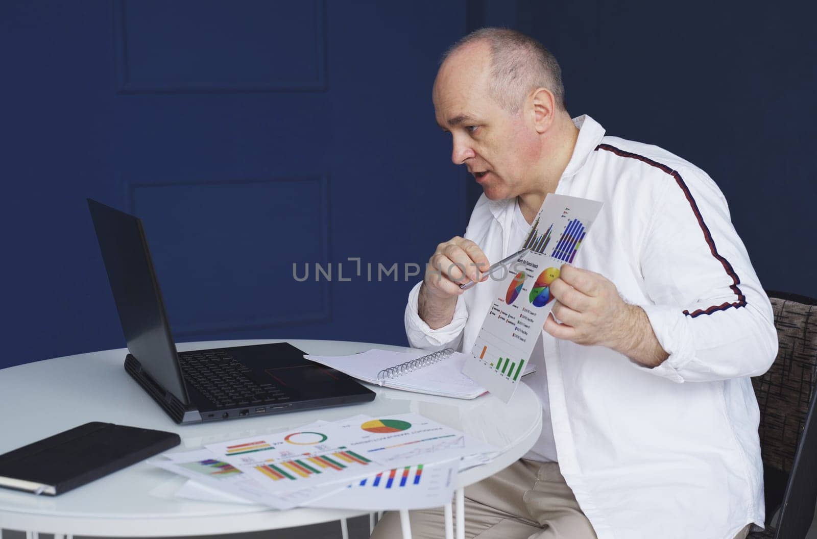 A man makes an online video call chat, sitting at home and talking into a laptop webcam, showing a document.