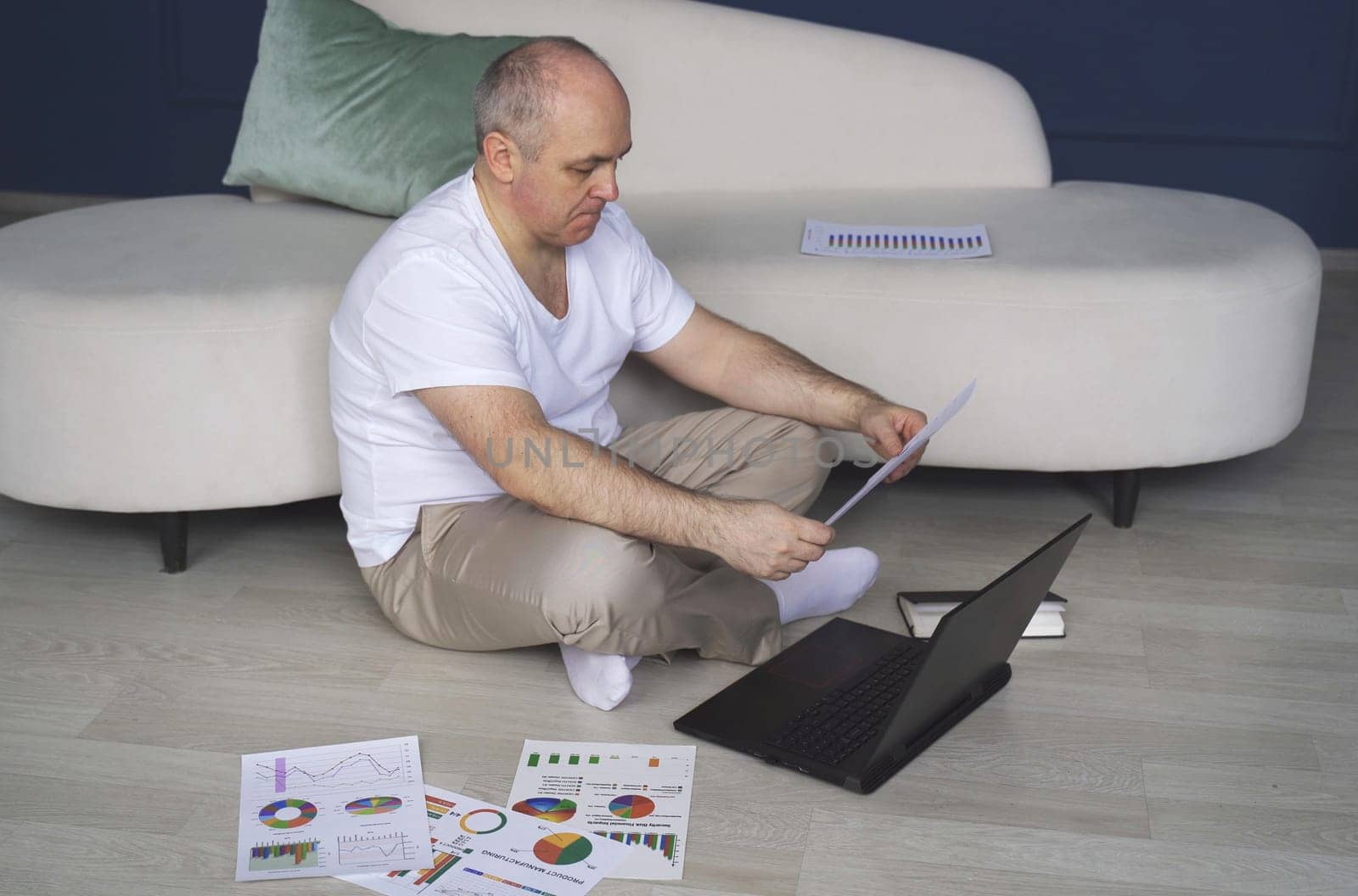 A man sits on the floor, works on laptops, studies reporting documents, charts and graphs.