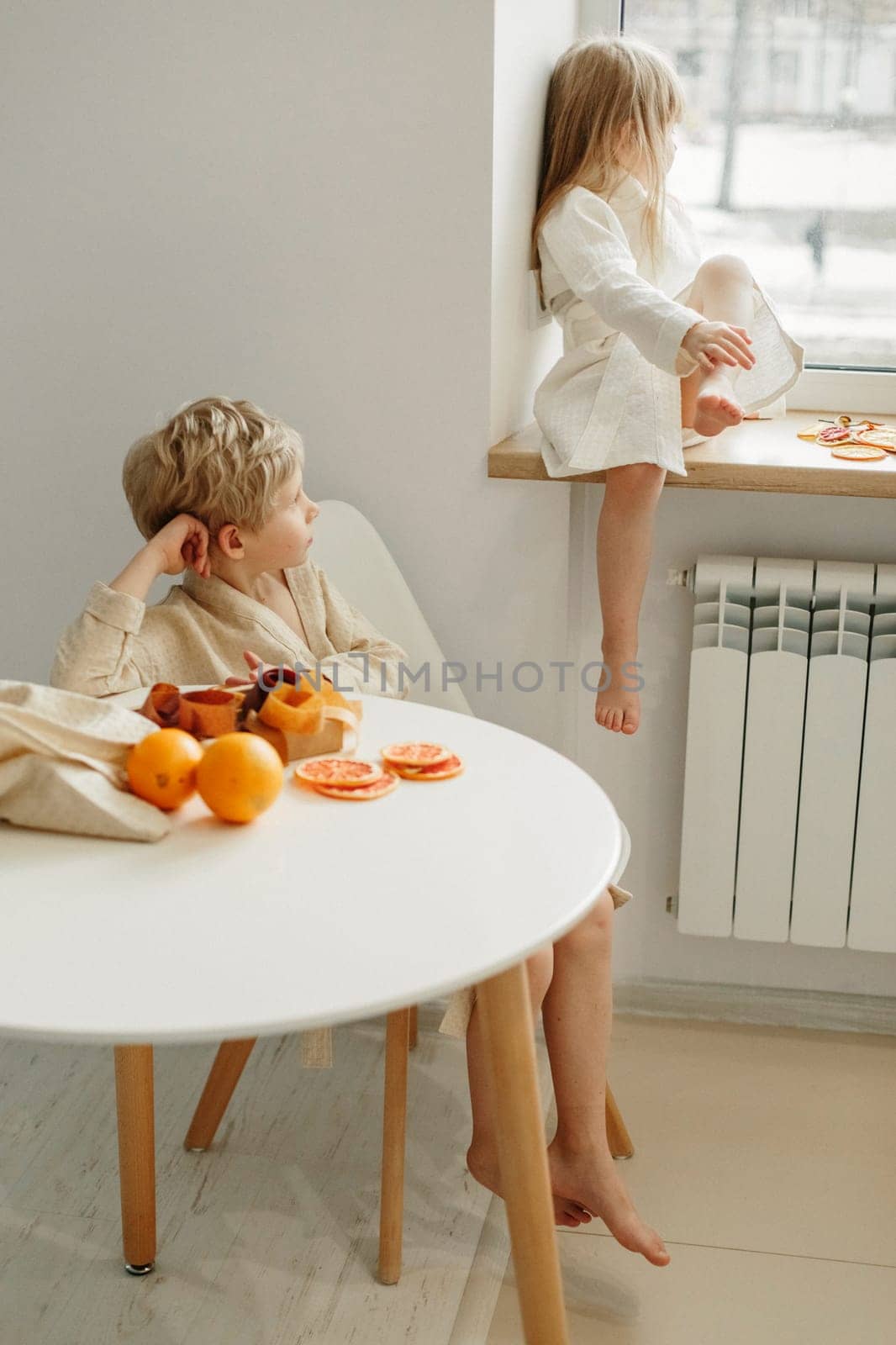 A boy and a girl in bathrobes are eating candied fruit and marmalade in the kitchen. The boy looks at the girl, the girl sits on the windowsill.