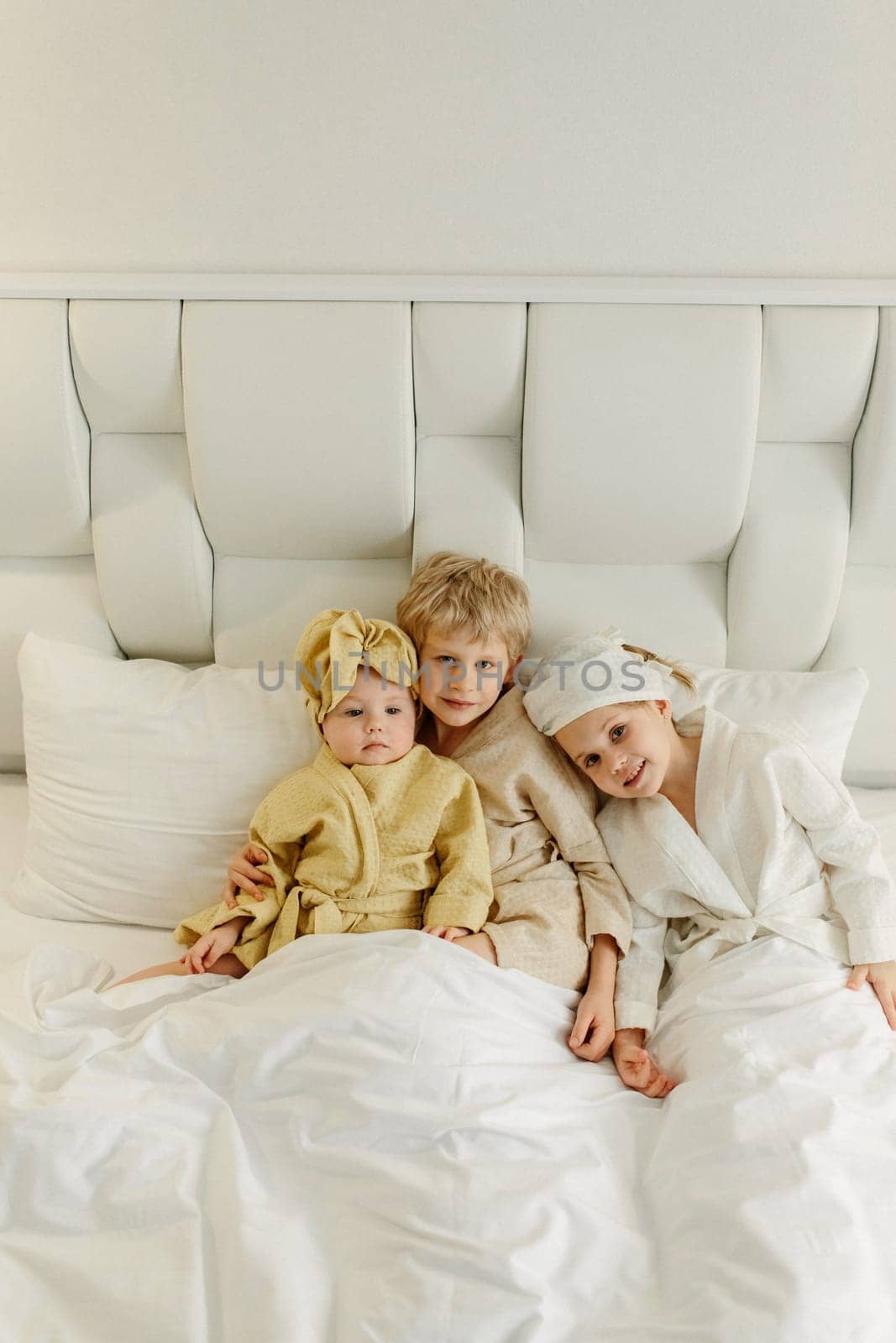 Children lie in a white bed in bathrobes after taking a bath, smiling and looking at the camera. View from above.