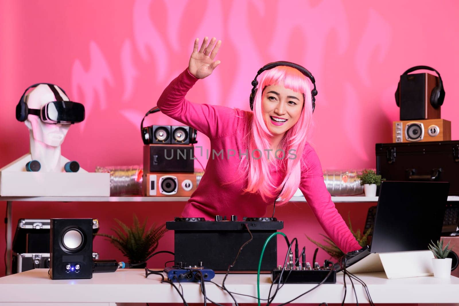Artist with pink hair and blouse having fun while mixing eletronic music with techno using mixer console, standing at dj table in club at night. Musical performer enjoying performing