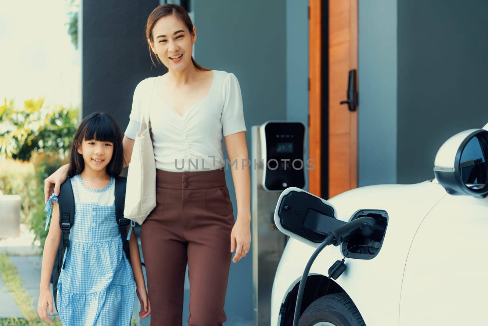 Progressive lifestyle of mother and daughter who have just returned from school in an electric vehicle that is being charged at home. Electric vehicle powered by sustainable clean energy.