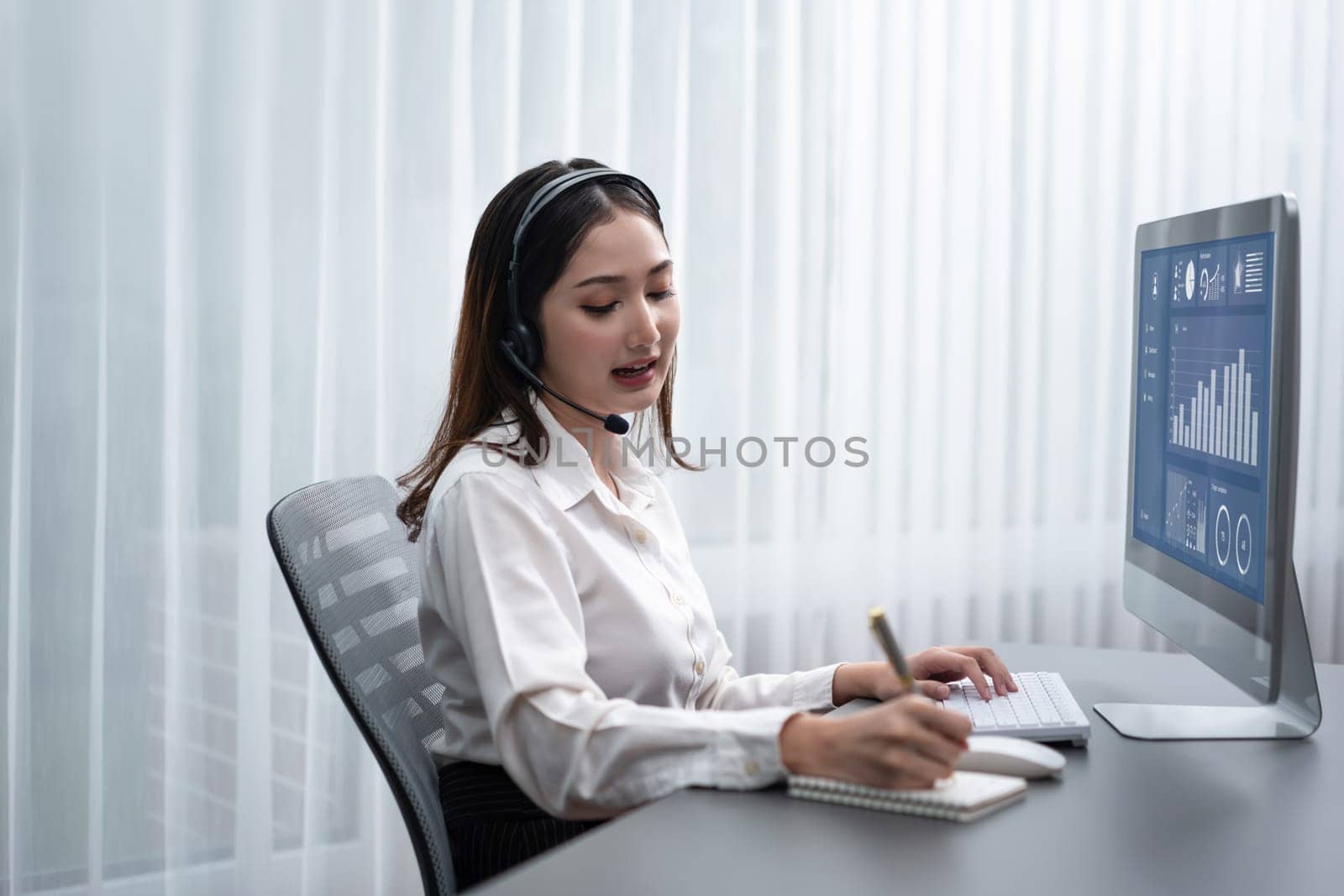 Enthusiastic asian call center with headset and microphone working on her laptop by biancoblue