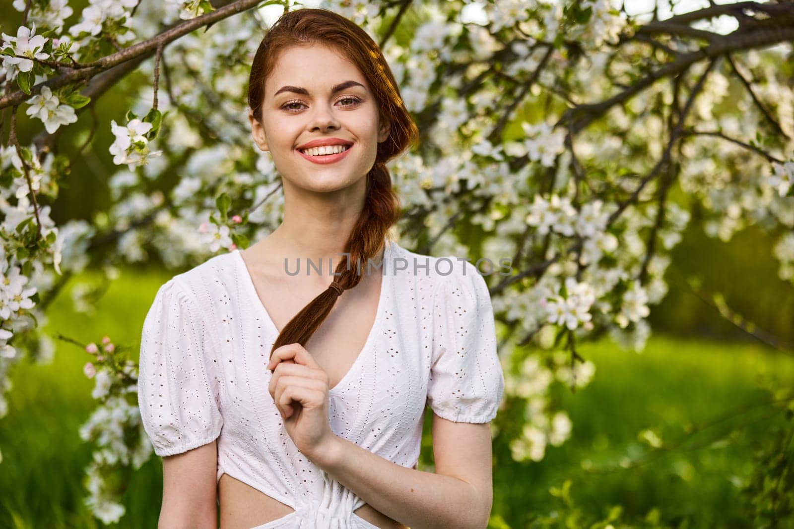 portrait of a joyful woman in a light dress against the background of a flowering tree, holding herself by her braid by Vichizh