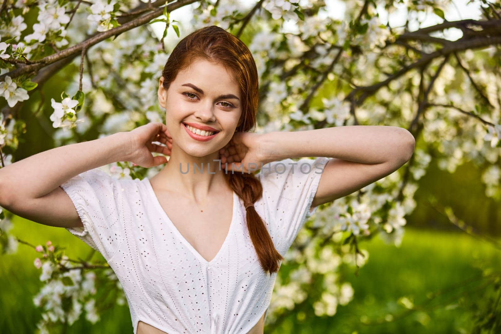 portrait of a joyful woman in a light dress against the background of a flowering tree, raising her hands to her head. High quality photo