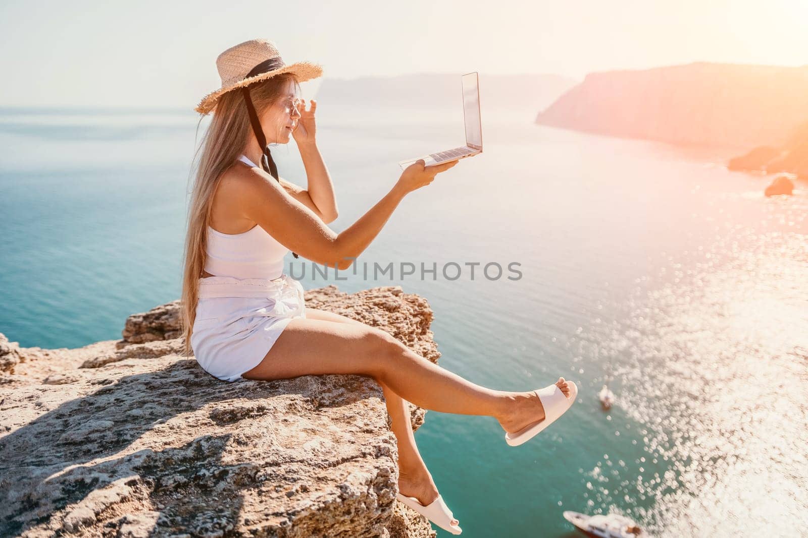 Working remotely on seashore. Happy successful woman female freelancer in straw hat working on laptop by the sea at sunset. Freelance, remote work on vacation