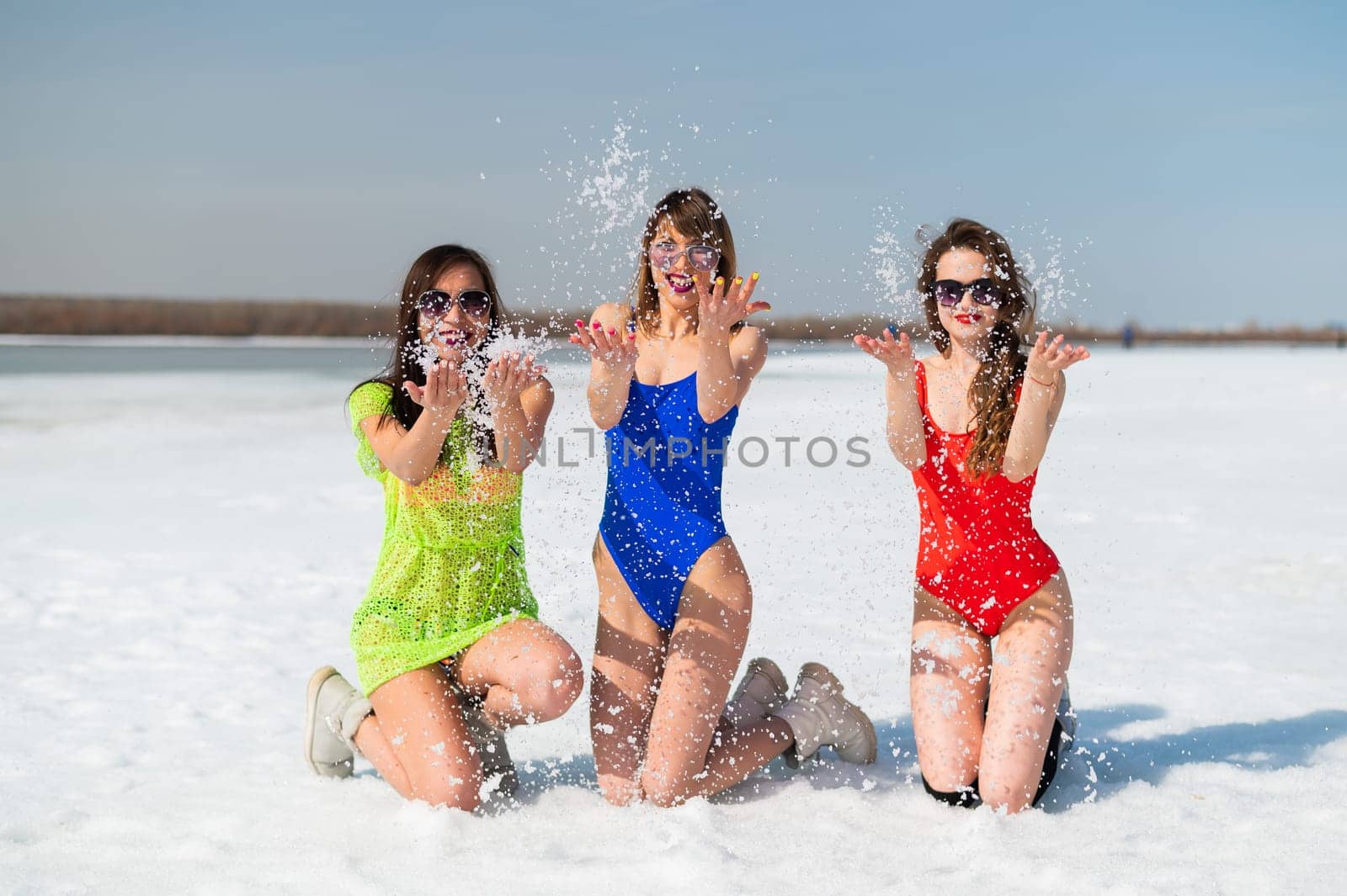 Three girlfriends in swimsuits are relaxing on a snow-covered beach. Hot girls posing in bikinis outdoors in winter