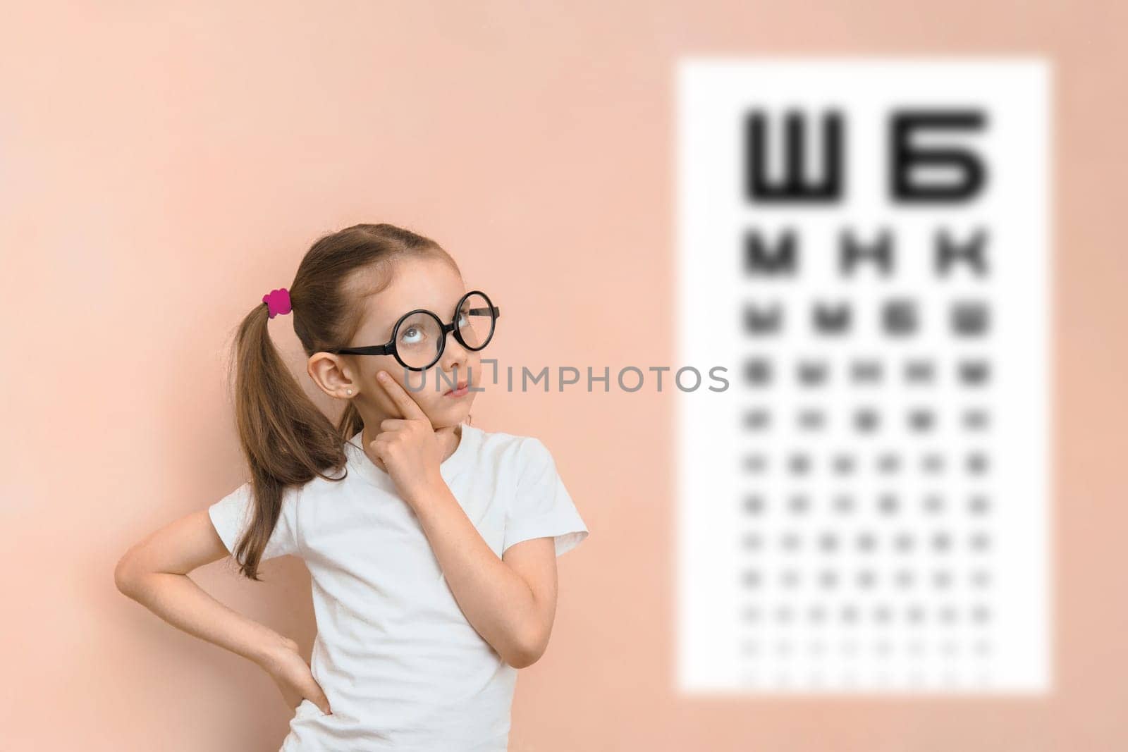 Thoughtful smart girl of elementary school age in round glasses against the background of a blurred sivtsev table for checking visual acuity.