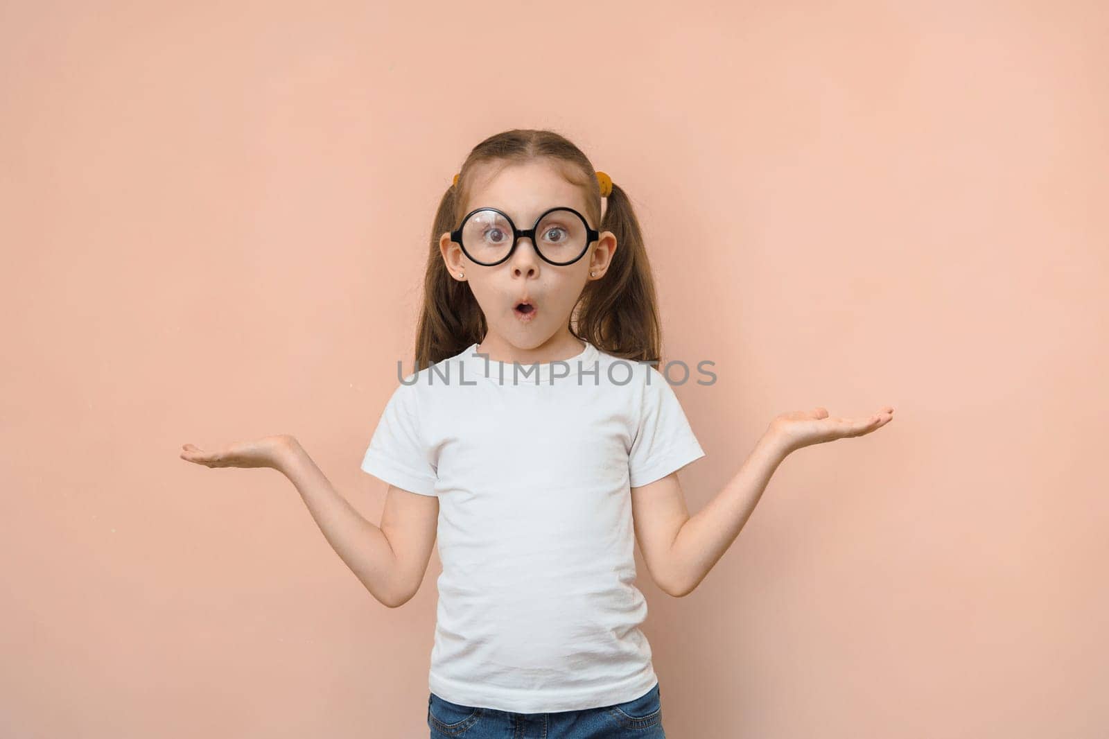 Cute funny little caucasian girl in round glasses can not choose what is in the empty space looking at the camera on a pink background.