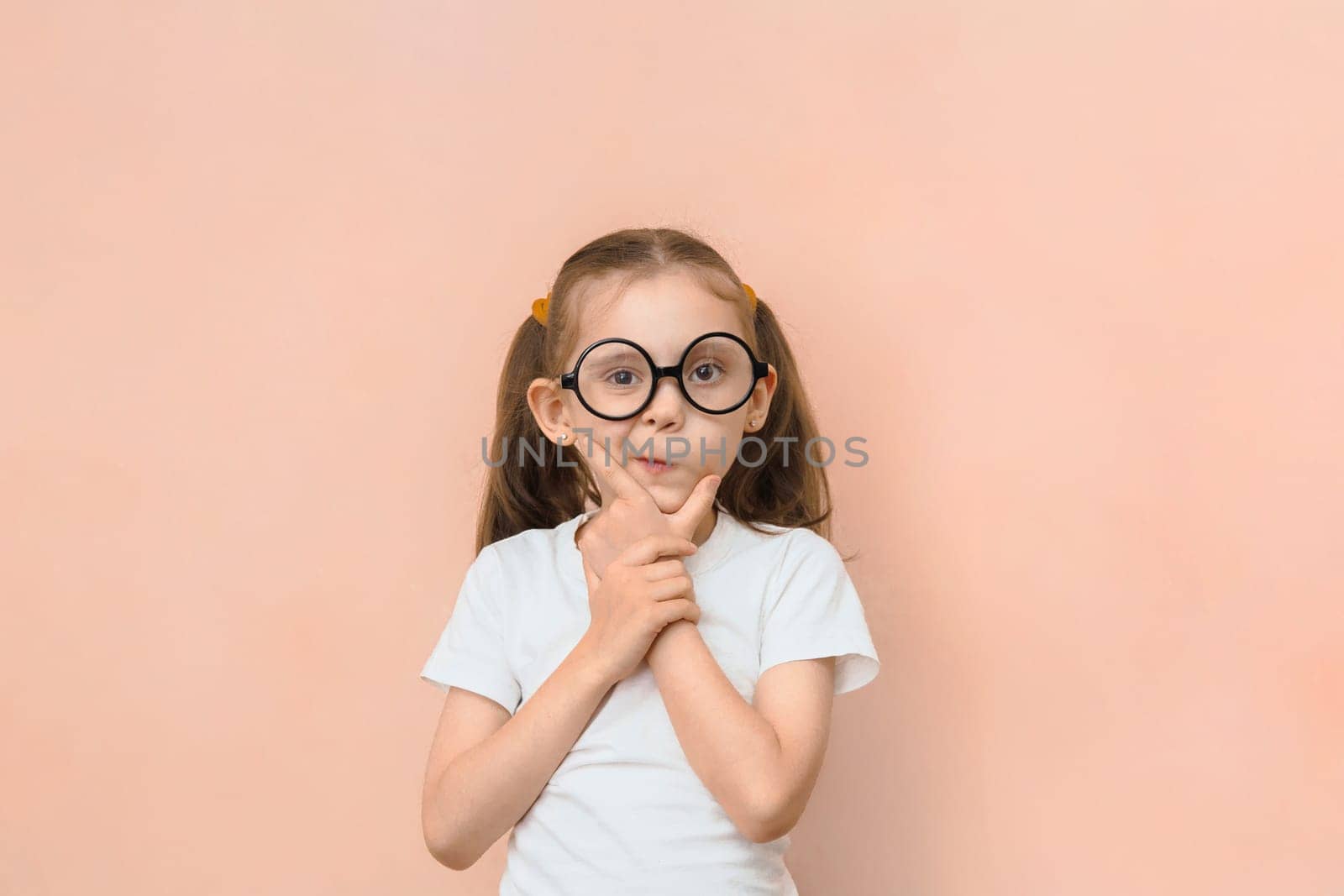 Cute funny little caucasian girl in round glasses and hands near her face is thinking something while looking at the camera on a pink background with copy space.