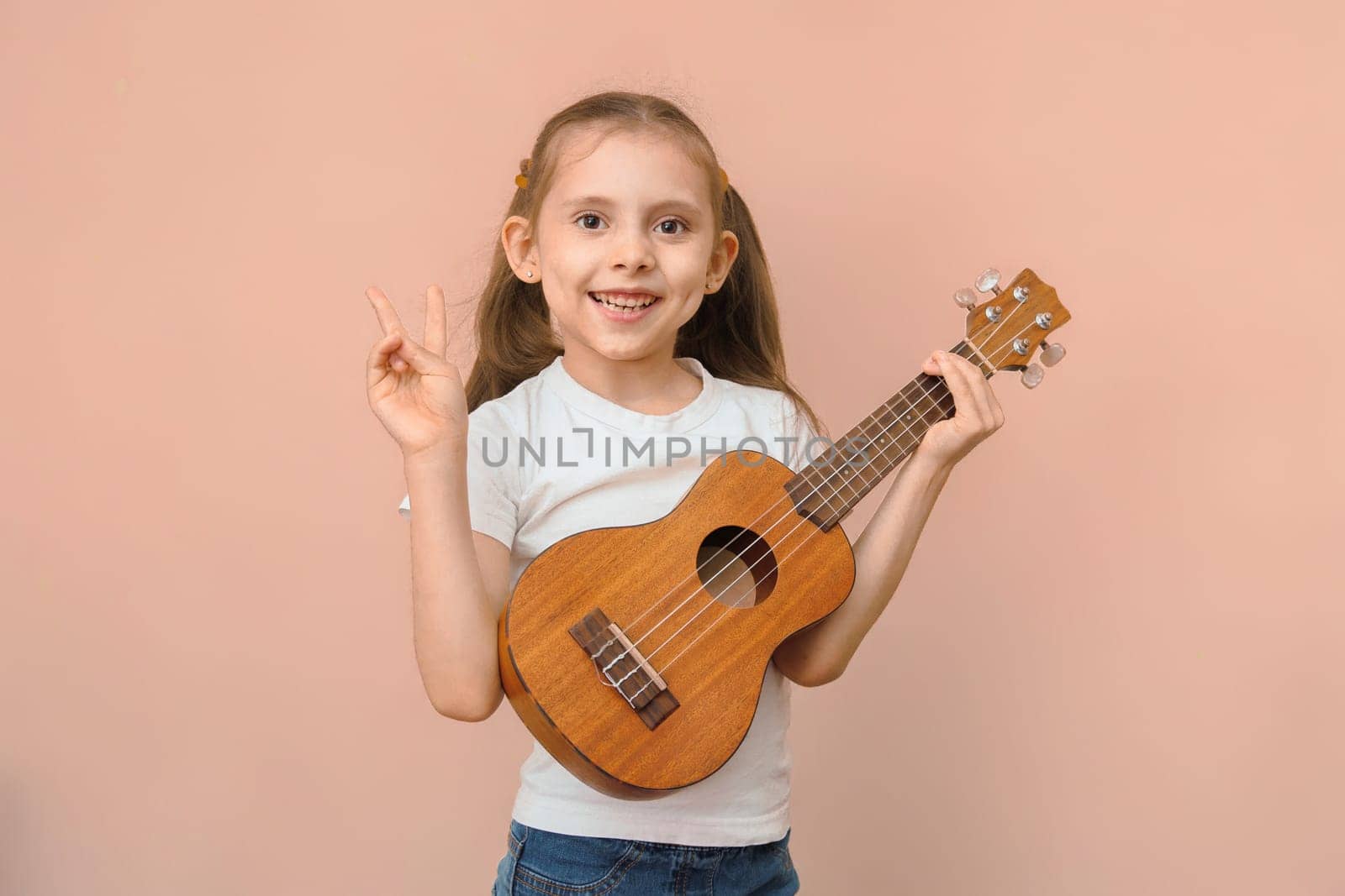 Cute caucasian baby girl in white t-shirt with ukulele shows peace gesture looks at camera on pink background.