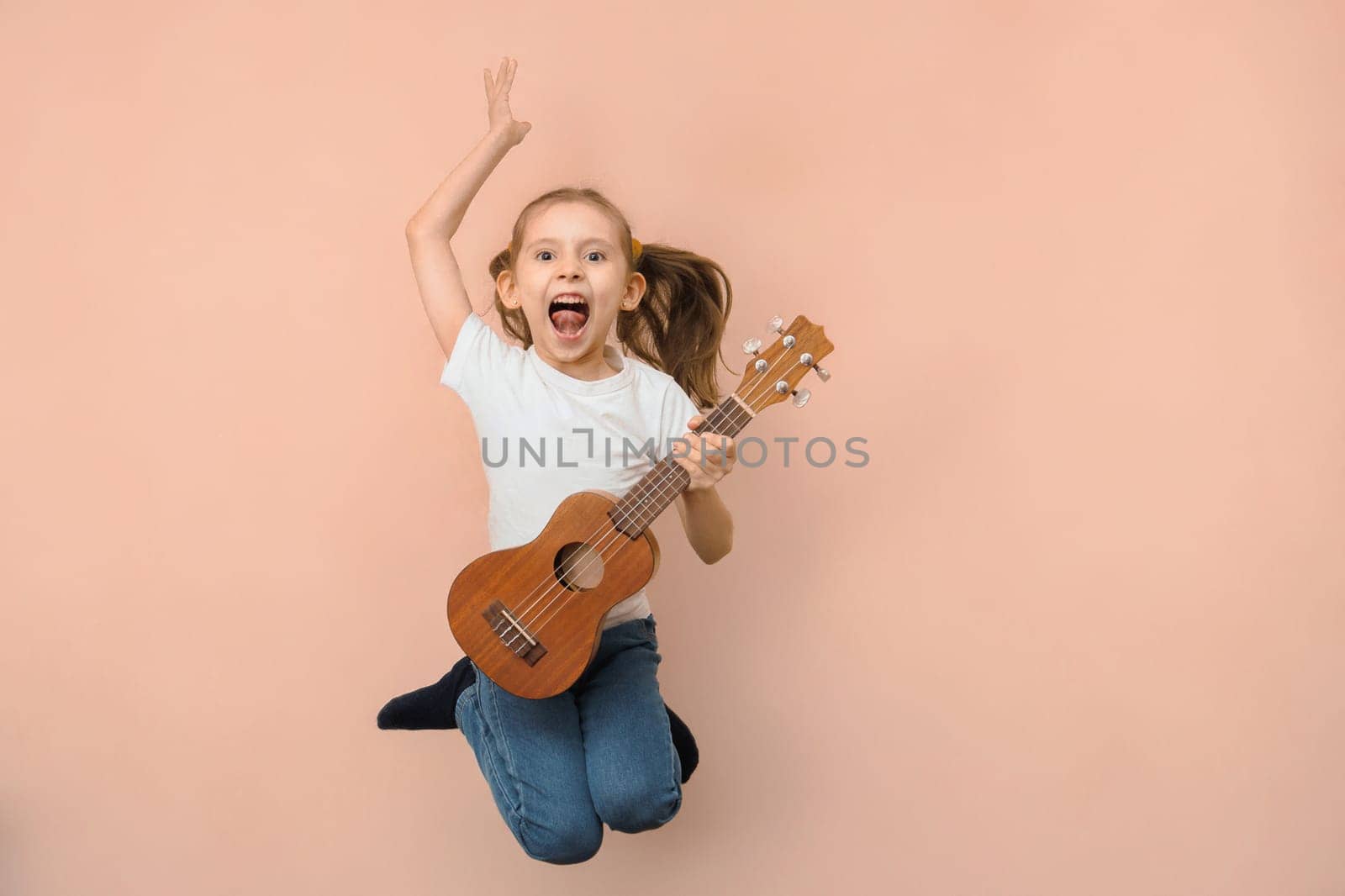 Emotional little jumping girl playing guitar like rock star on pink background.