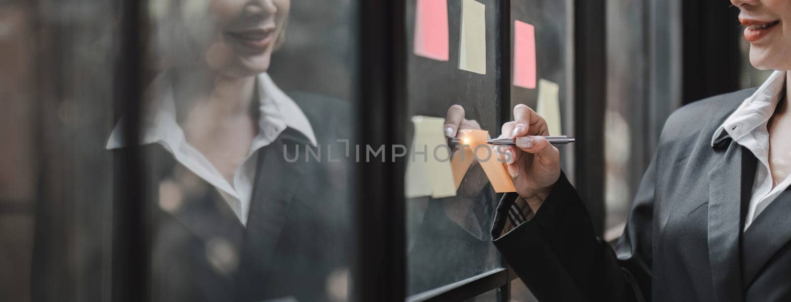 Young businesswoman creative team using post it notes in glass wall to writing strategy business plan to development grow to success...