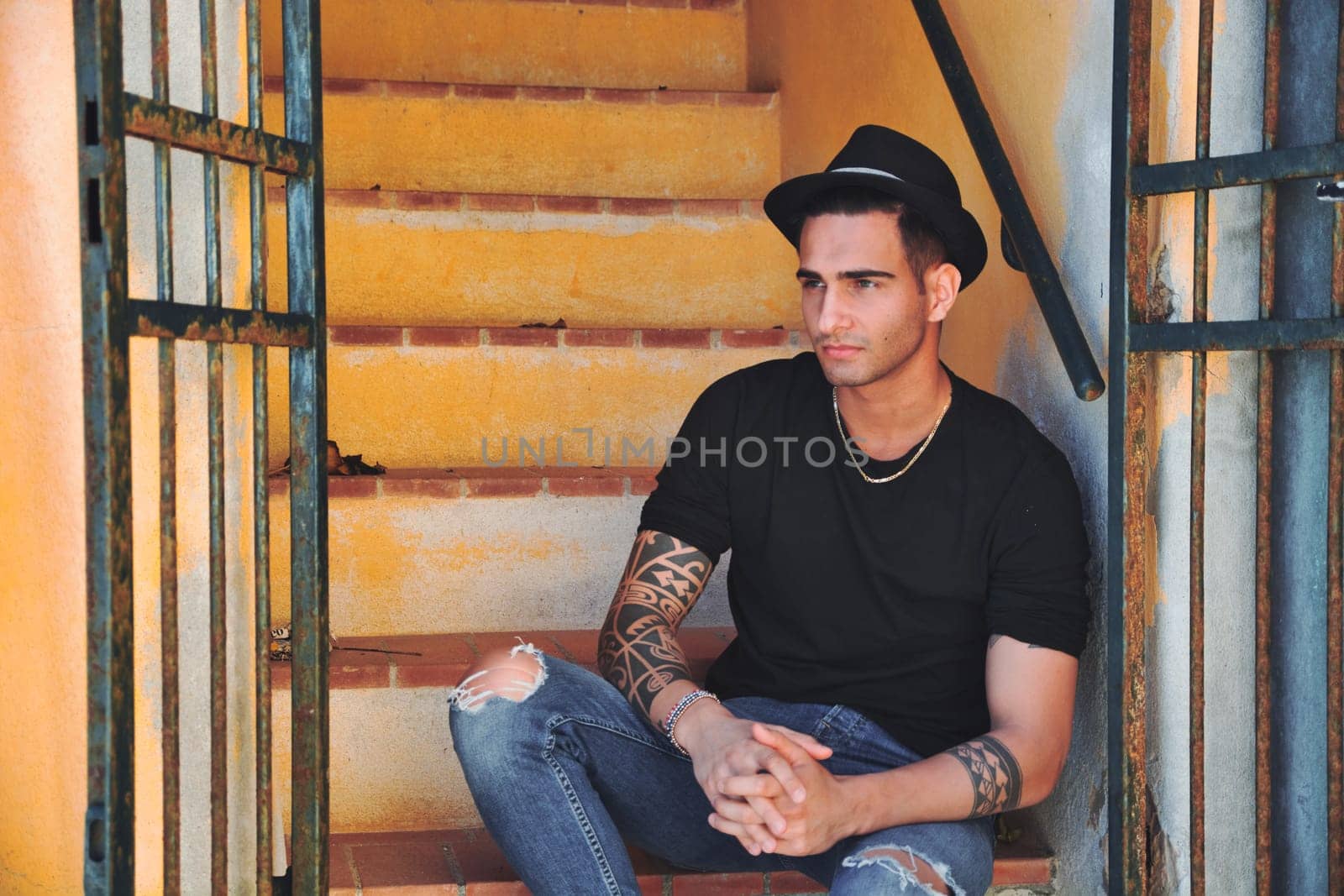 Portrait of handsome man with tattoo on arms sitting on cement stairs while leaning on wall, looking away to a side