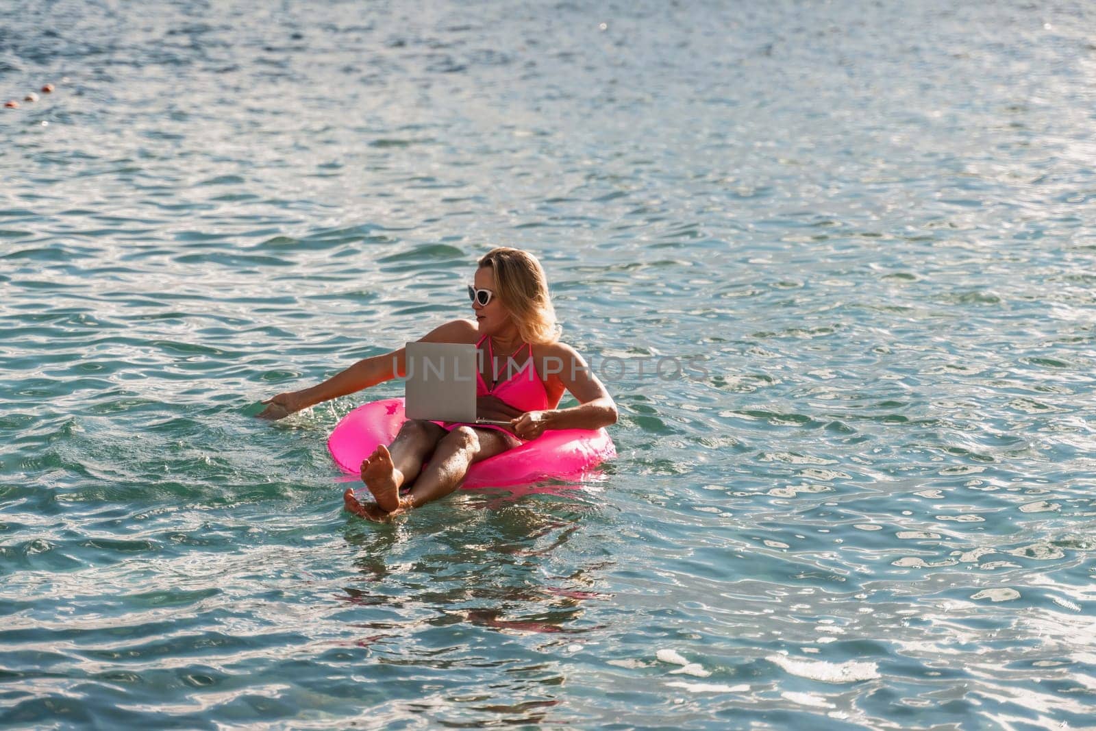 Woman works on laptop in sea. Freelancer, blond woman in sunglases floating on an inflatable big pink donut with a laptop in the sea at sunset. People summer vacation rest lifestyle concept. by Matiunina