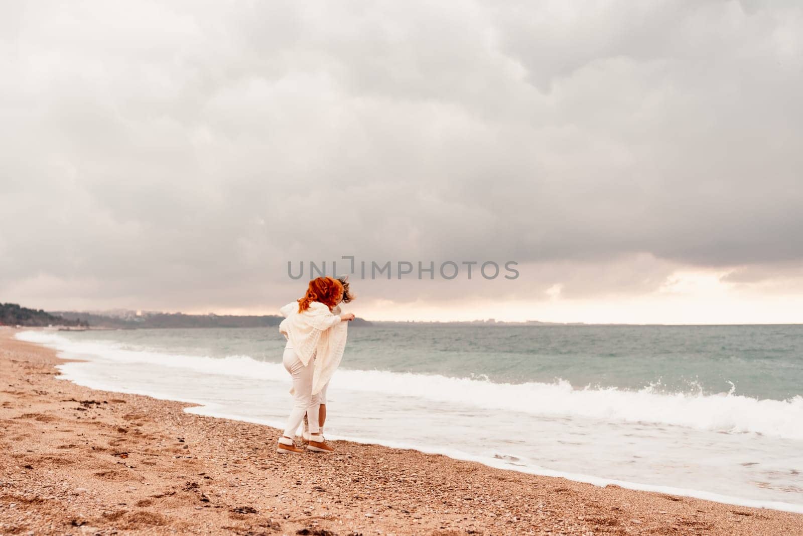 Women sea walk friendship spring. Two girlfriends, redhead and blonde, middle-aged walk along the sandy beach of the sea, dressed in white clothes. Against the backdrop of a cloudy sky and the winter sea. Weekend concept. by Matiunina