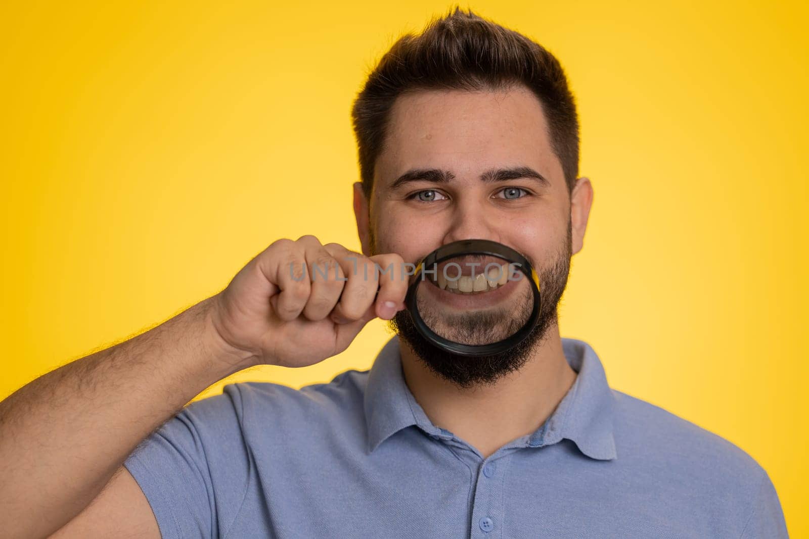 Caucasian man holding magnifier glass on healthy white teeth, looking at camera with happy expression, showing funny silly face smiling mouth. Handsome young guy isolated on yellow studio background