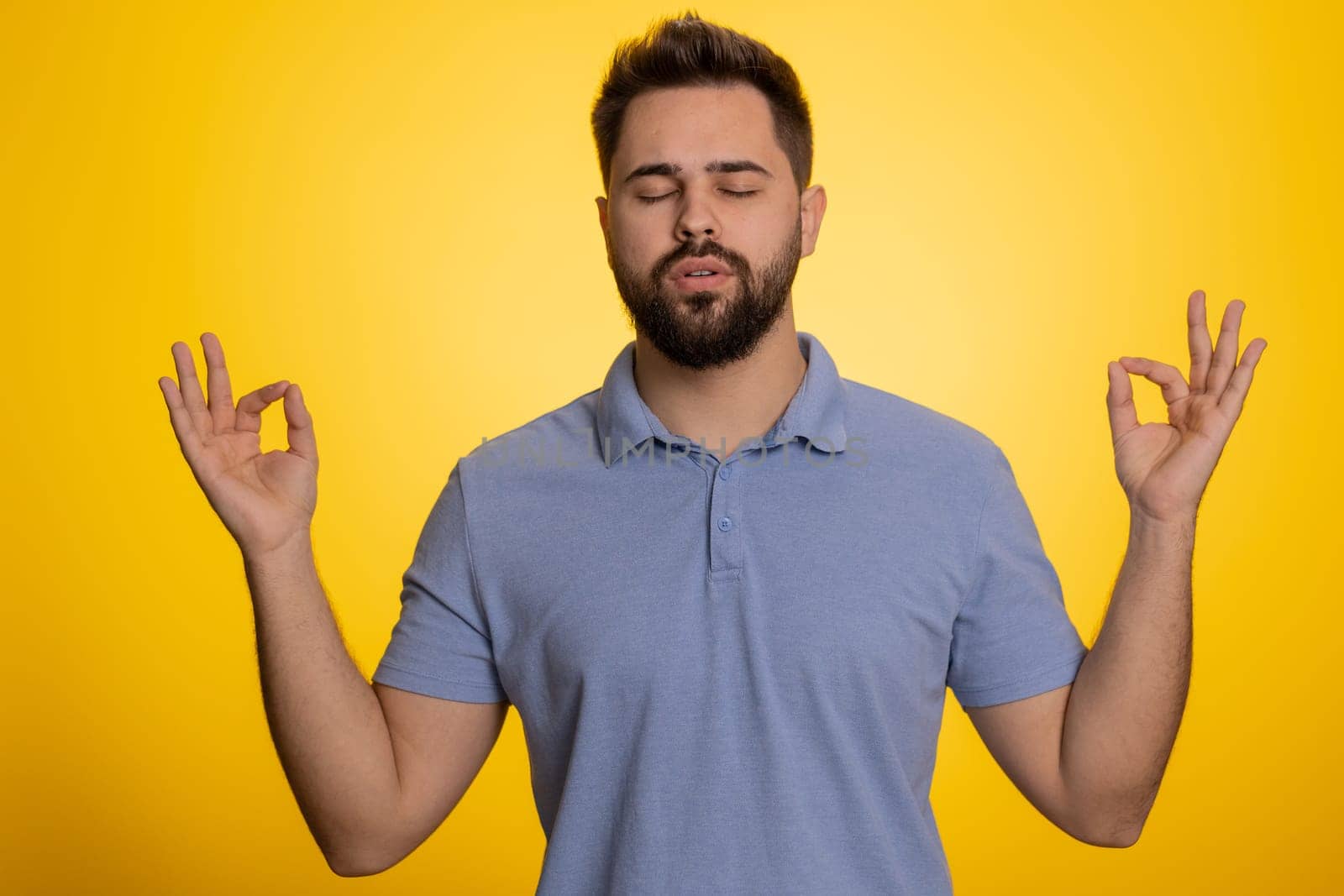 Keep calm down, relax, inner balance. Young caucasian man breathes deeply with mudra gesture, eyes closed, meditating with concentrated thoughts, peaceful mind. Guy isolated on yellow background