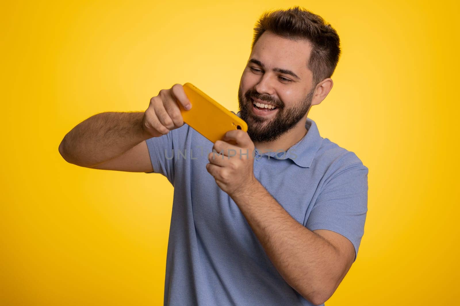 Worried funny addicted caucasian man enthusiastically playing racing video games on mobile phone. Young adult guy using smartphone gadget app with drive simulator isolated on yellow studio background