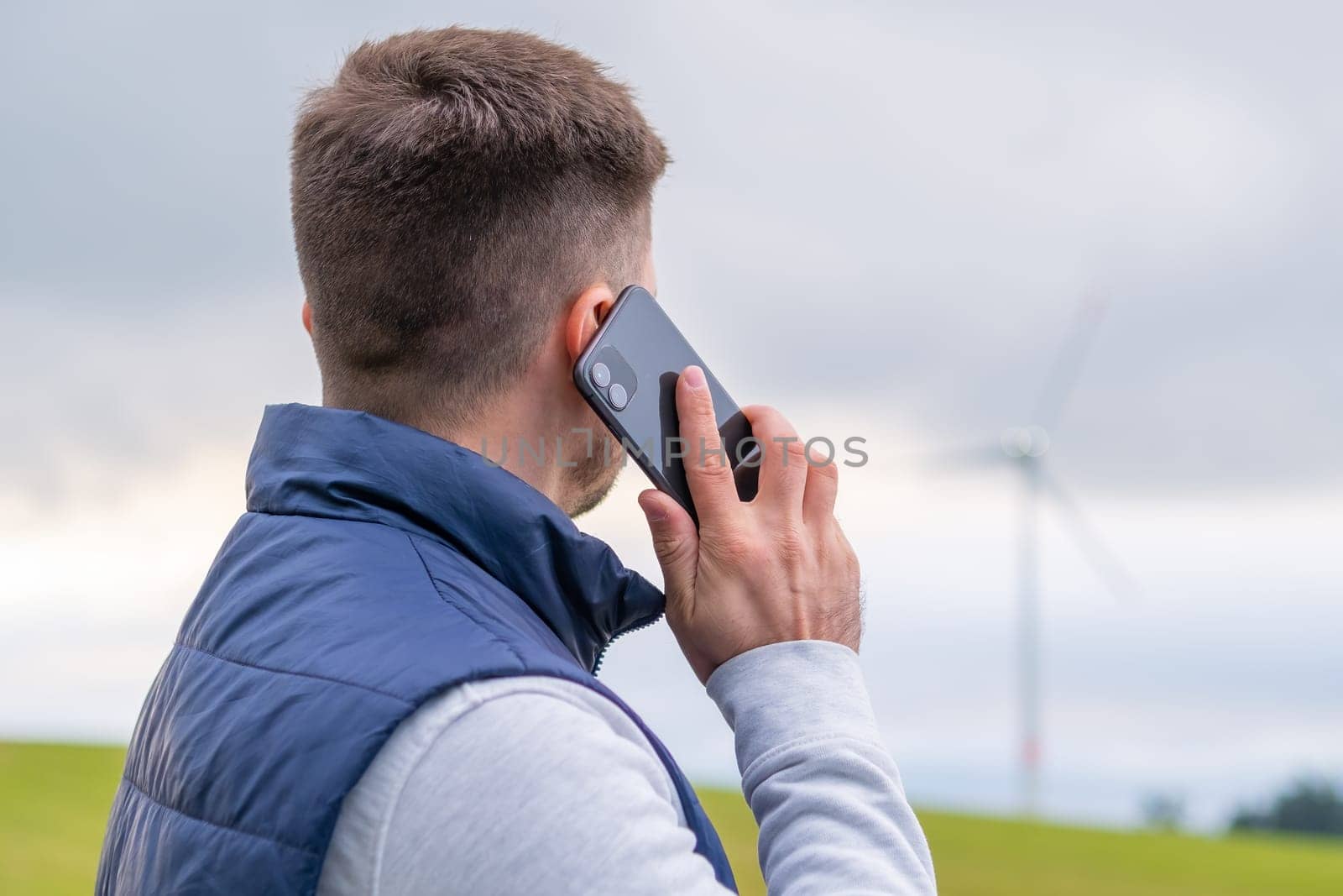 Man engineer reports about functioning wind turbine via smartphone looking at windmill. Wind turbine produces green energy against grey sky