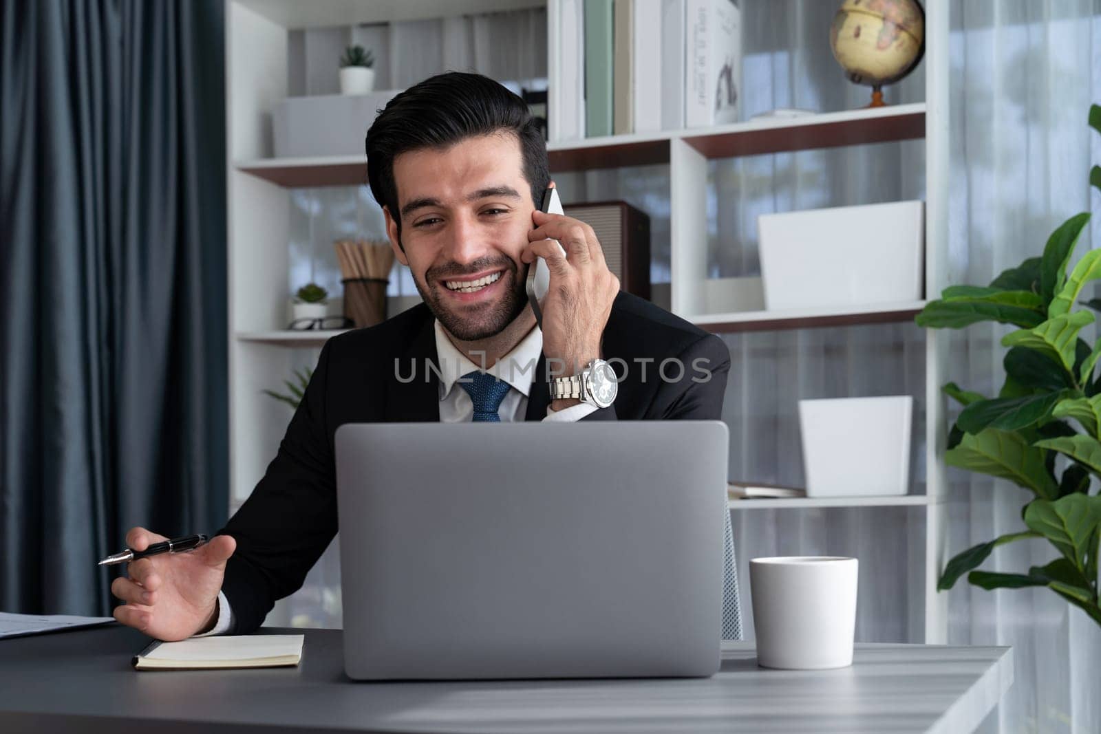 Diligent businessman busy talking on the phone call with clients while working with laptop in his office as concept of modern hardworking office worker lifestyle with mobile phone. Fervent