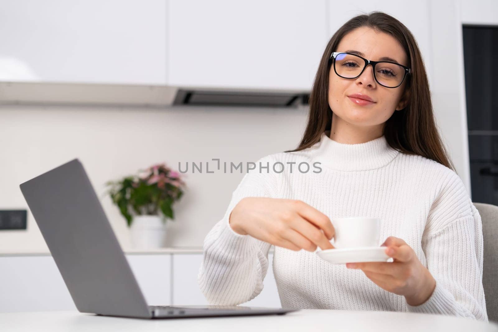 Portrait of a young beautiful woman holding a coffee cup and sitting in the kitchen near the laptop. Freelancer has a break