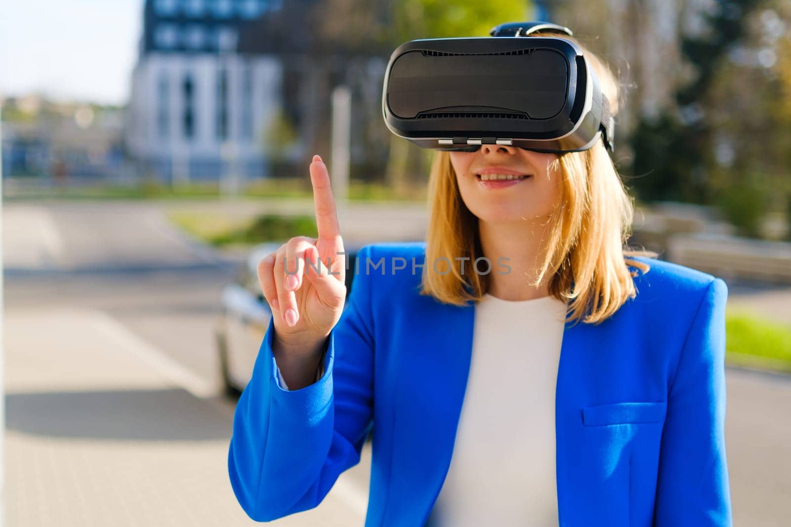 A happy woman in the suit is standing outside and gestures in augmented reality in 3D VR goggles.