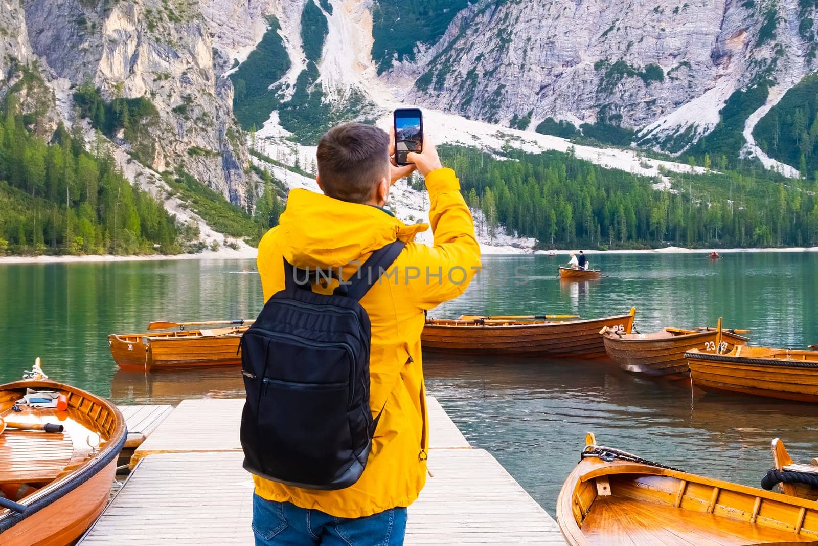 Tourist taking photo of wooden boats and Dolomites on the lake Braies. Man wearing yellow jacket and holding a black backpack visiting Dolomites Alps, Italy.