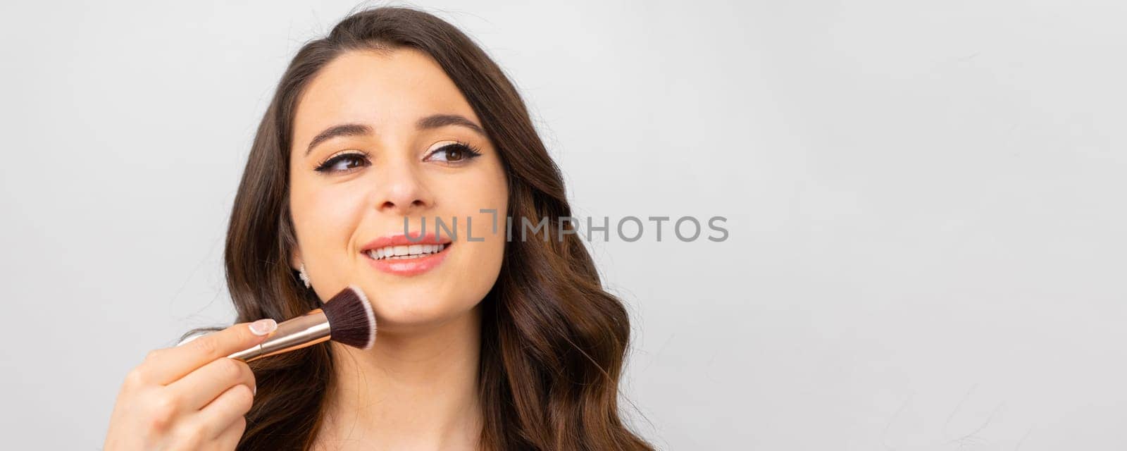 Young woman with perfect skin holding makeup brush near her face on the grey background with copy space