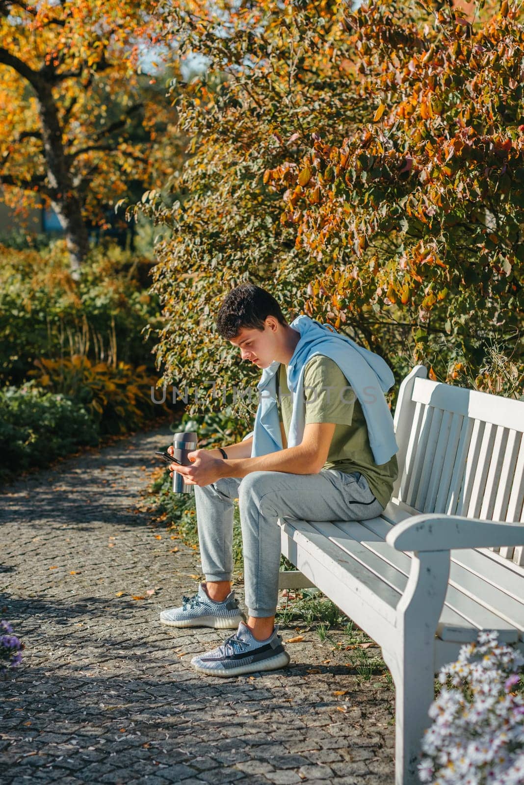 A Teenager Sits On A Bench In The Autumn Park Drinks Coffee From A Thermo Mug And Looks Into A Phone. Portrait Of Handsome Cheerful Guy Sitting On Bench Fresh Air Using Device Browsing Media Smm Drinking Latte Urban Outside Outdoor by Andrii_Ko