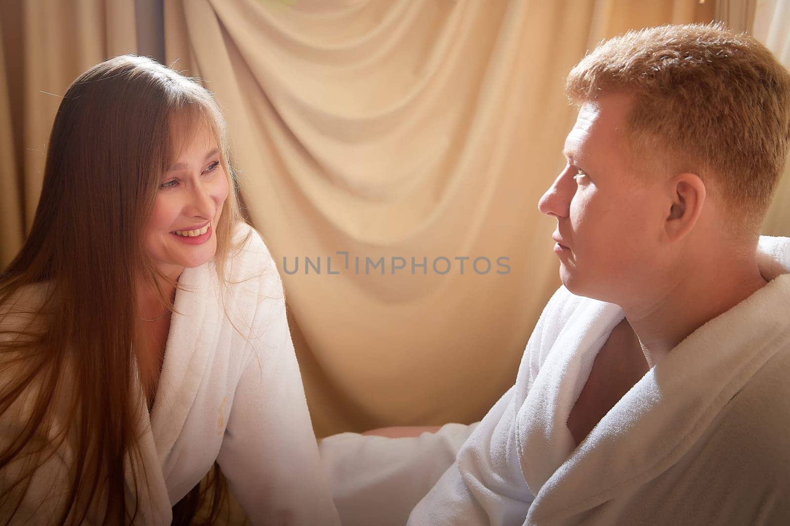 An adult couple of newlyweds in in white robes in a hotel room after the wedding. A guy and a girl, a man and a woman in beautiful room. The concept of hugs, love and care