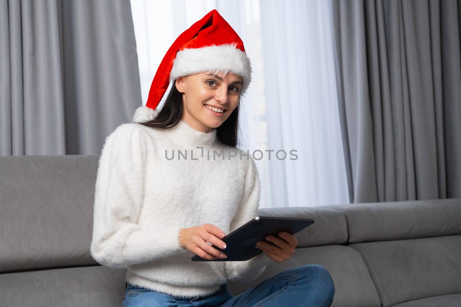 Christmas portrait of a beautiful girl wearing a Santa Claus hat and sitting on the sofa with a tablet in her hand in a modern home interior. Ordering of gifts via the internet on Christmas or New Year.