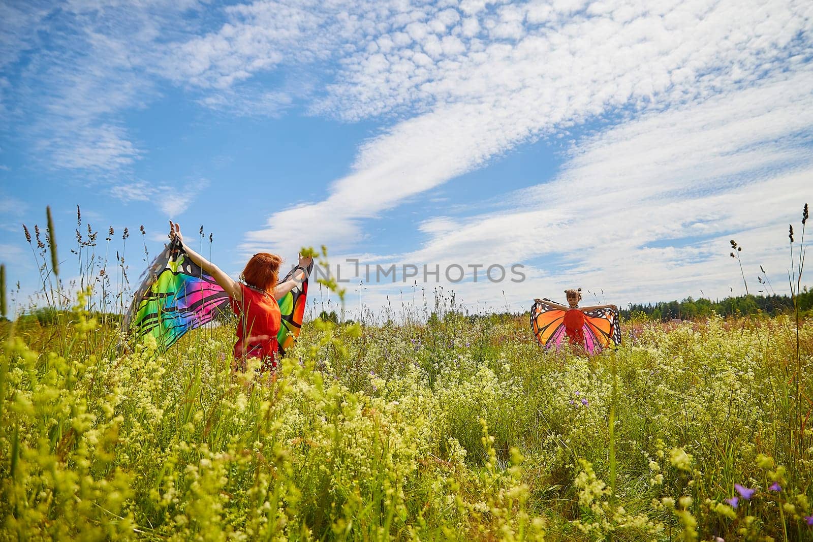 Happy female family with red haired mother and daughter with bright butterfly wings having fun on green and yellow meadow full of grass and flowers in sunny summer day. Concept family love