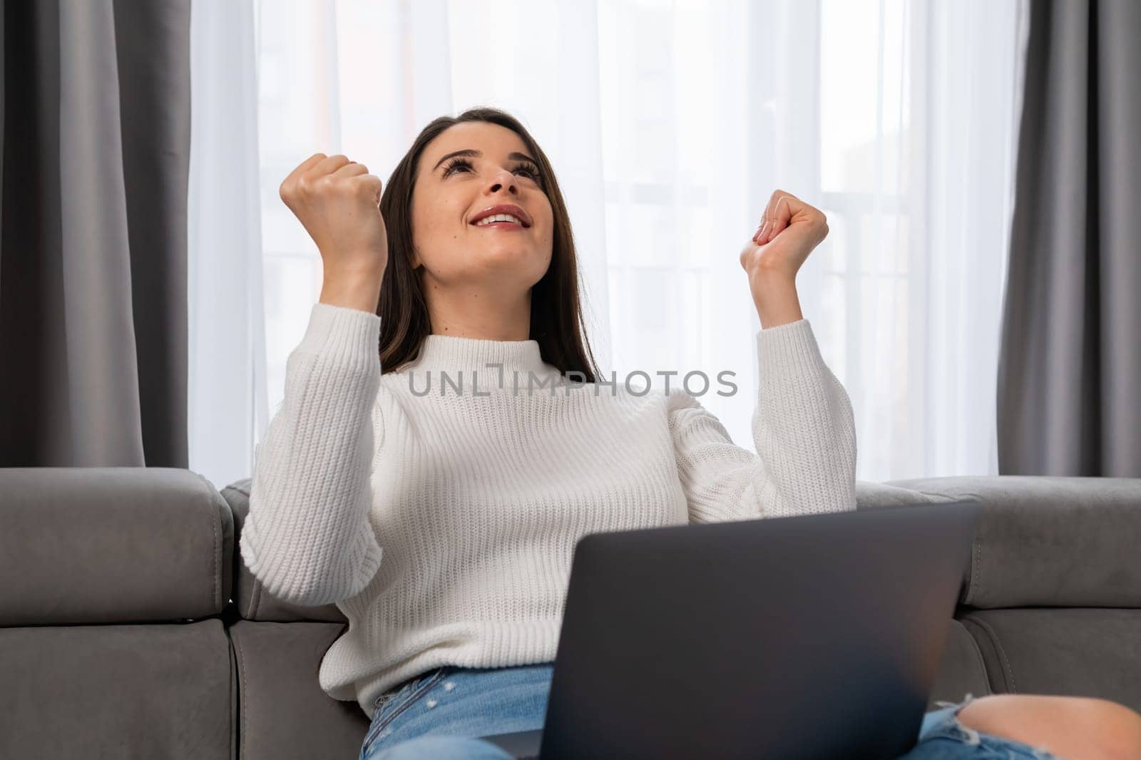 An excited young woman rejoices in internet victory holding a laptop on her lap in a cozy living room.