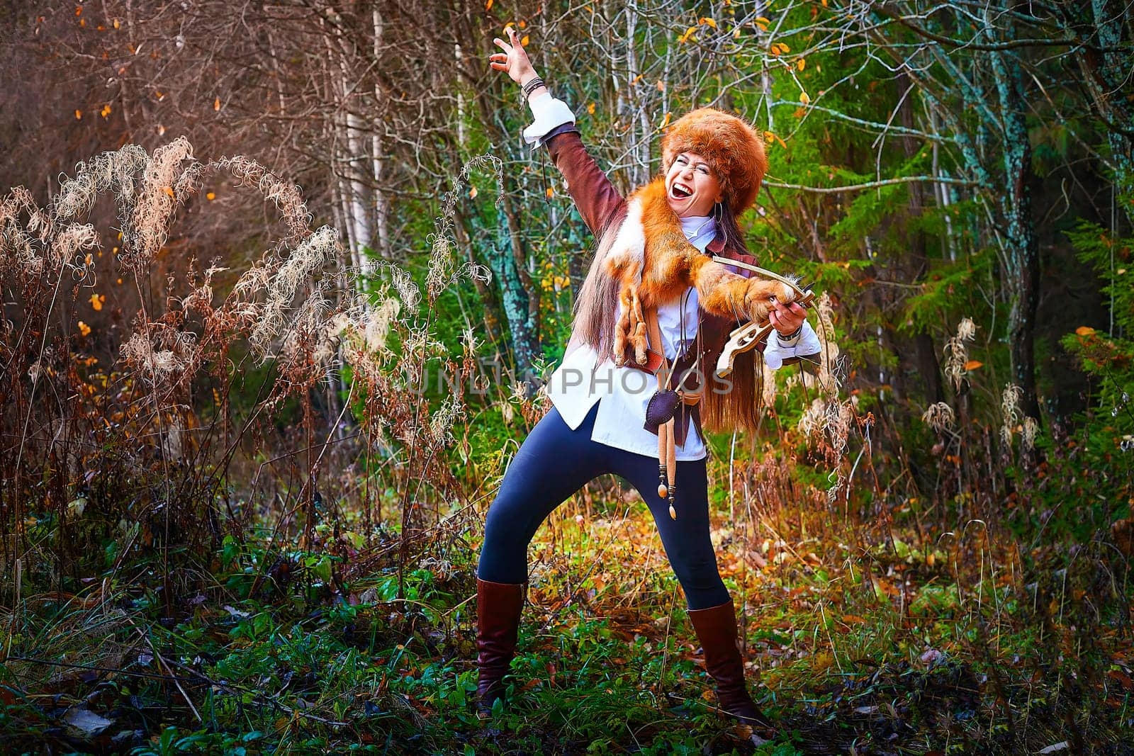 Girl in a leather jacket, a big red fox fur hat and with the skin of a fox killed on the hunt in the forest in autumn. A female model poses as fabulous royal huntress on nature hunt at photo shoot