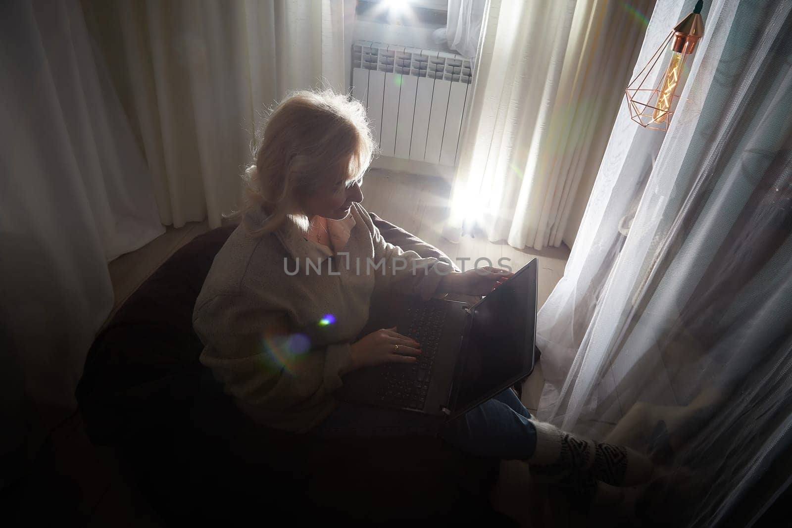 Silhouette and shadow of Adult mature woman of 40-60 years old with laptop, notebook, computer in warm sweater in dark calm cozy evening atmosphere room. Interior with curtains and small linght by keleny