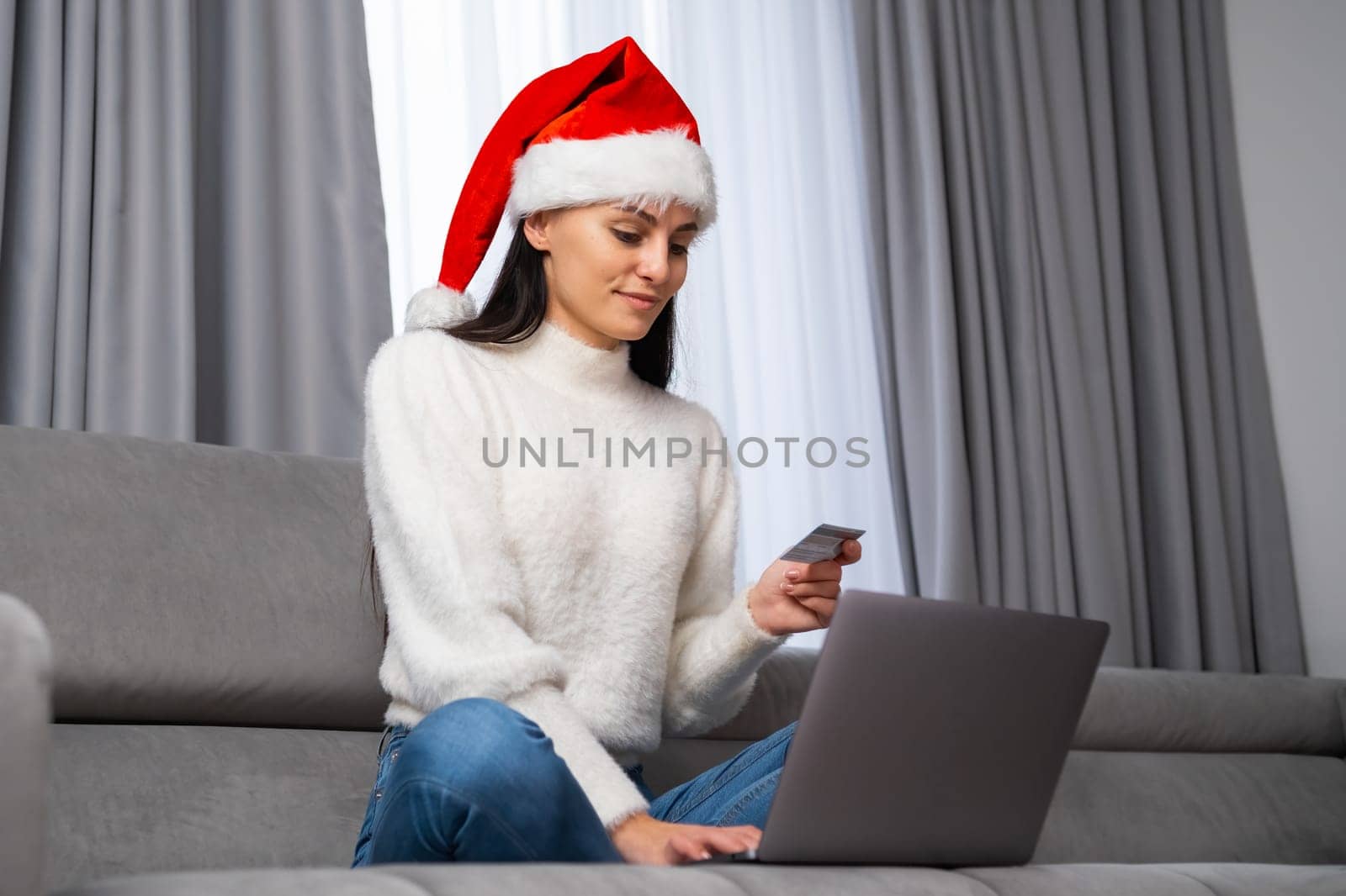 A beautiful young woman in a Santa Claus hat uses a credit card and makes payment using a laptop while sitting in the living room.