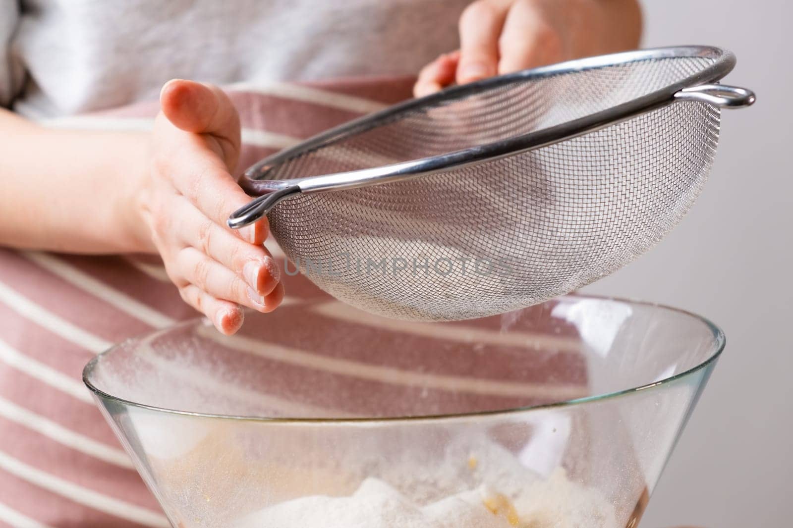 Close up woman sifts wheat flour through a sieve into mixing bowl for dough preparation for cake and bakery on the black background. Knead the dough for homemade pastry.