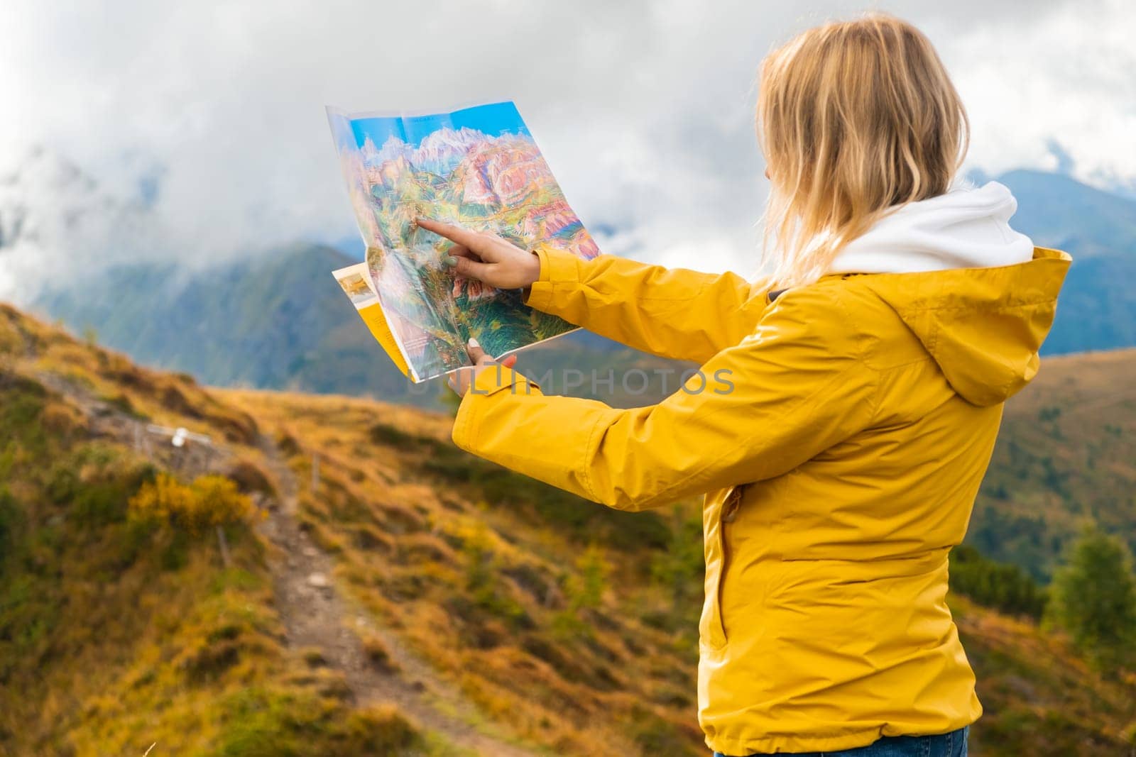 Traveling woman in yellow sport jacket standing with a map in front of mountains. Wanderlust concept.
