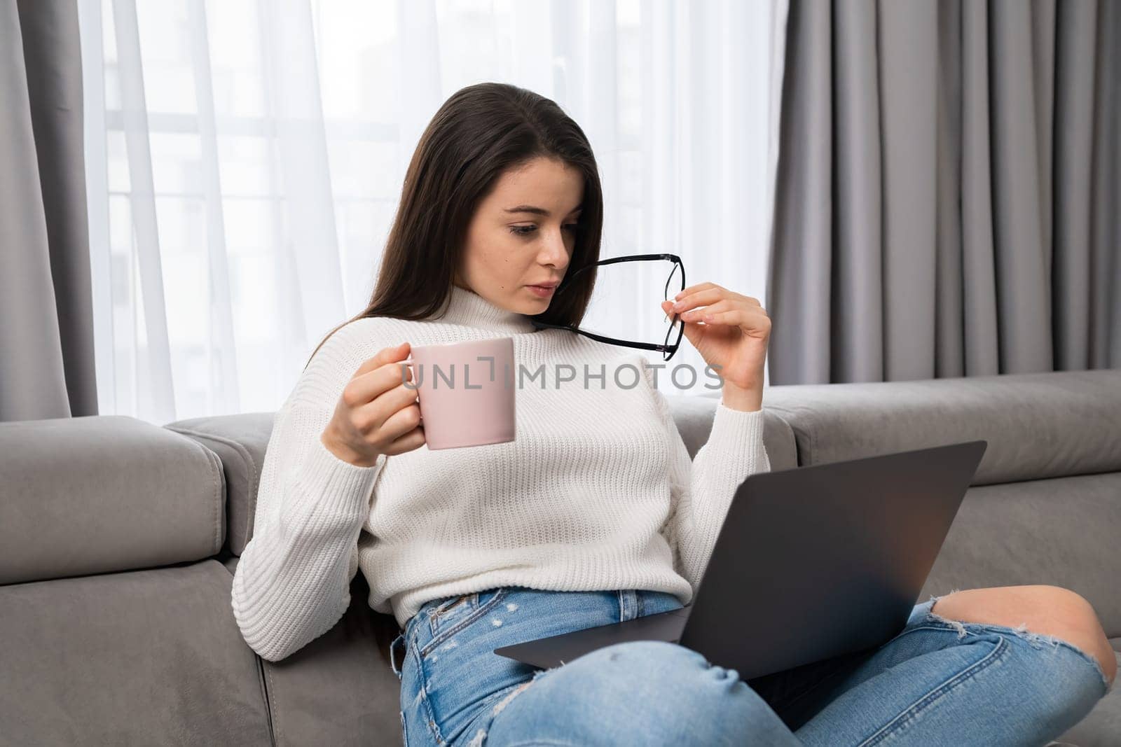 Satisfied woman checks catalogue of new clothes via Notebook holding glasses. Young lady customer rests on soft couch using modern laptop at home, holding cup of tea and glasses in the hands.