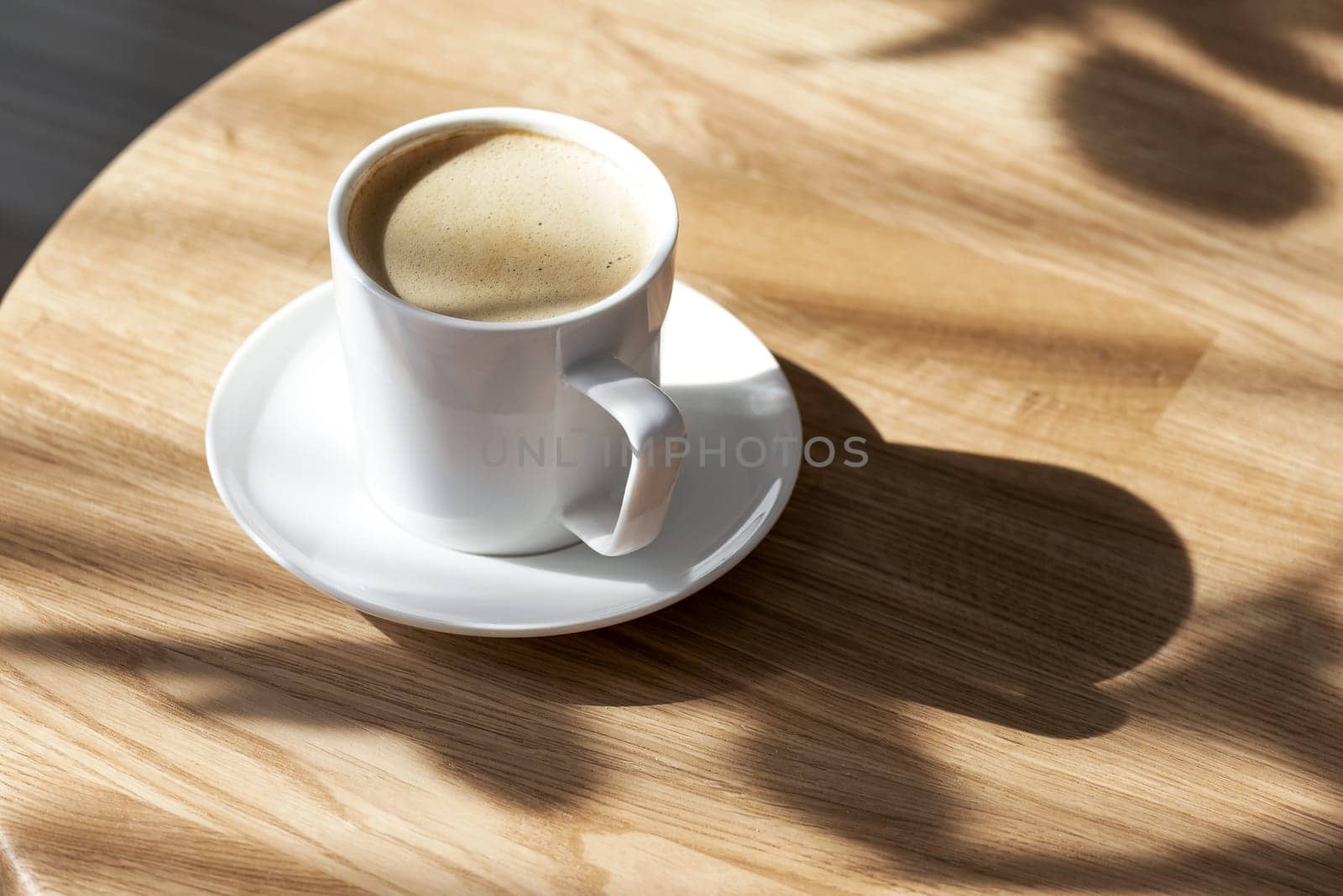 A white porcelain cup with coffee on a saucer stands on a small round table made of natural wood. Morning, coffee, hot drink. Hard light.