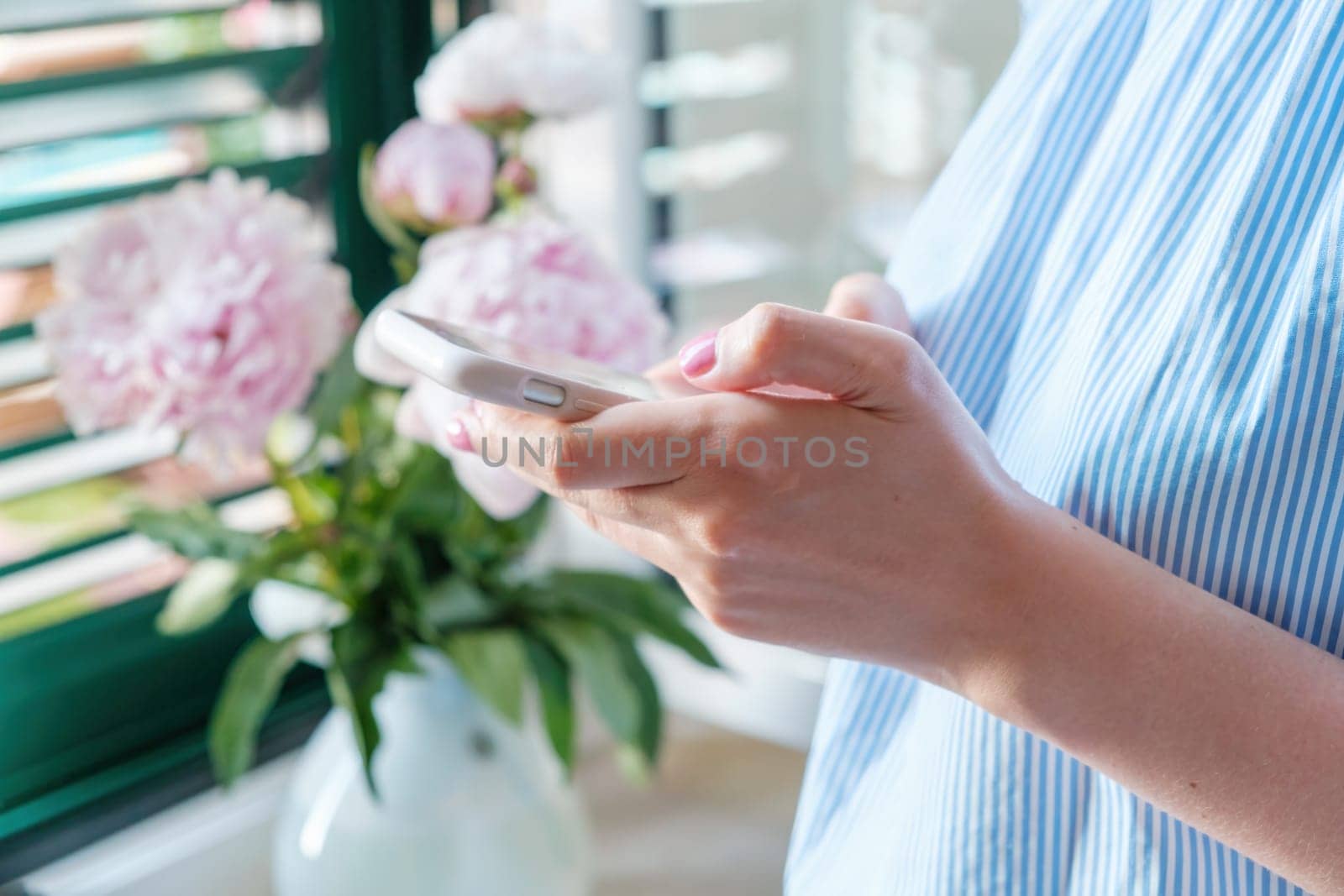 Young woman uses the phone on the background of beautiful pink peonies near the window. Romantic atmosphere at home