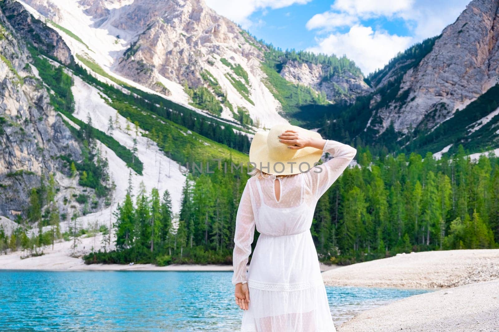 Back view of woman in white dress and hat on the shore of Lake Braies against mountains. Vacation and travel concept.