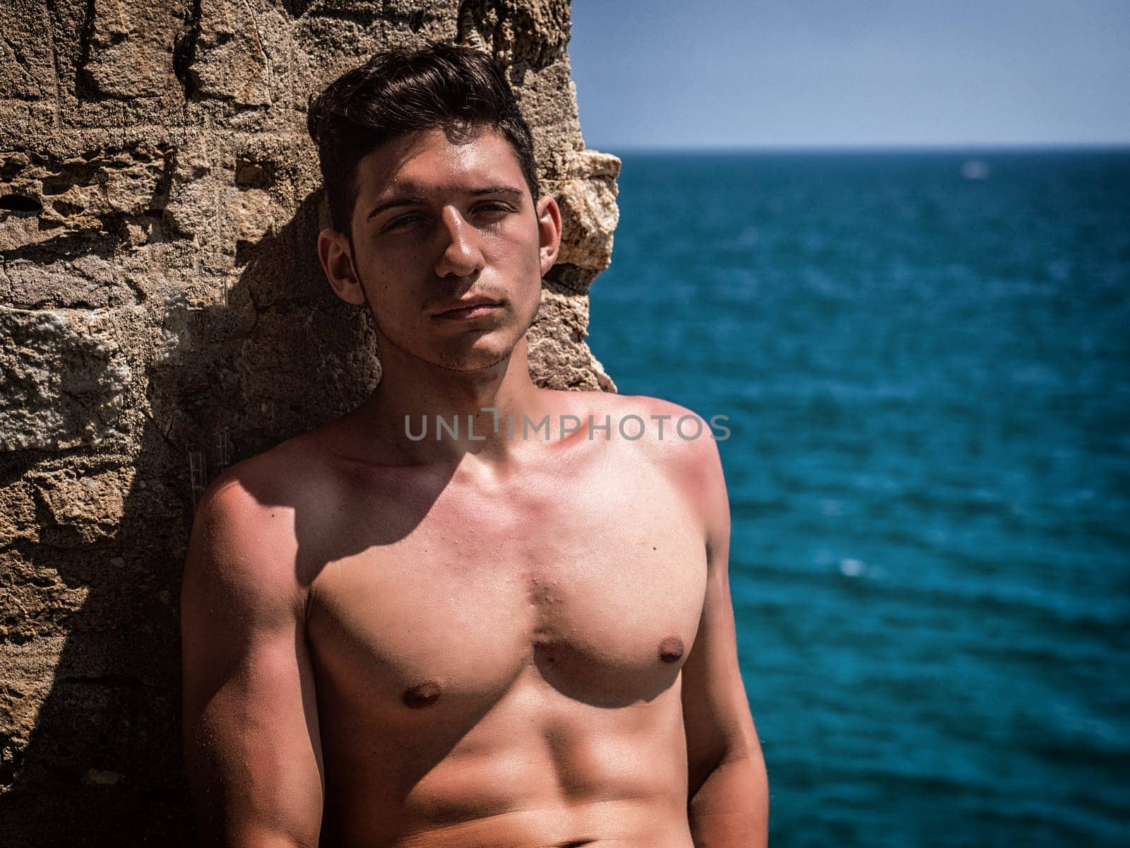 Handsome fit young man sitting by the sea, shirtless in a sunny day